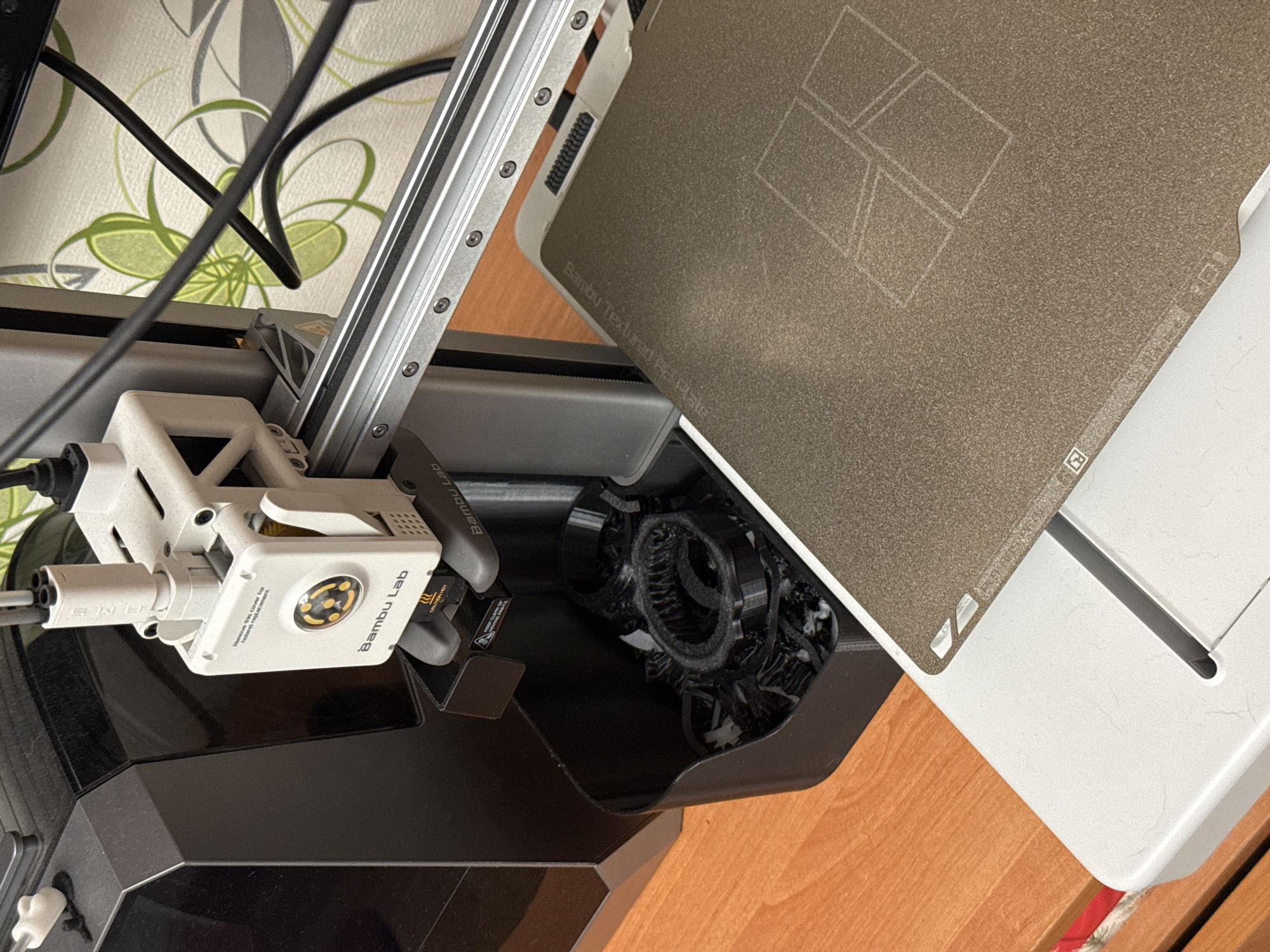 The image shows a close-up of a 3D printer. The printer's build plate is visible in the foreground, with a textured surface and a faint outline of printed squares. The print head assembly is mounted on a silver rail system, and cables run from it towards the back. To the left of the print head, a black bin is filled with discarded 3D printed parts. In the background, a floral patterned wallpaper is visible, and a wooden surface forms the base of the scene. There are no people or animals present in the image. The scene is indoors, likely in a workshop or a home setting given the materials and equipment. The overall impression is that of a functional, active 3D printing environment.