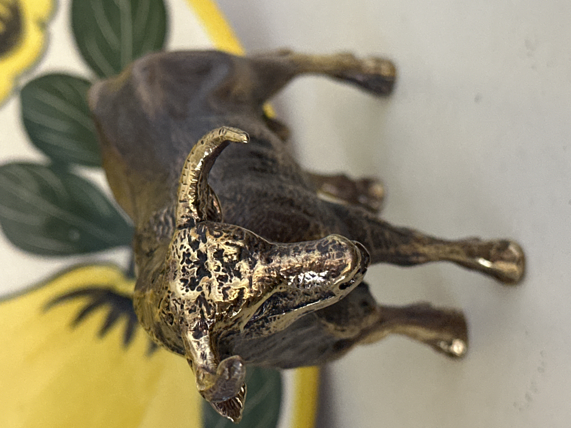 A close-up, shallow depth of field shot shows a small, metallic bull figurine with intricate detailing and a textured, aged bronze finish. The figurine is positioned on a plain, light-colored surface. In the background, out of focus, are elements of what appears to be a decorative object with large, stylized yellow and green leaf or petal shapes. The lighting is bright, casting highlights on the metallic surface of the bull, particularly on its horns, head, and legs. The image does not contain any identifiable features of the city of Nevinnomyssk, Russia, nor any people or discernible environmental context beyond the immediate tabletop or surface.