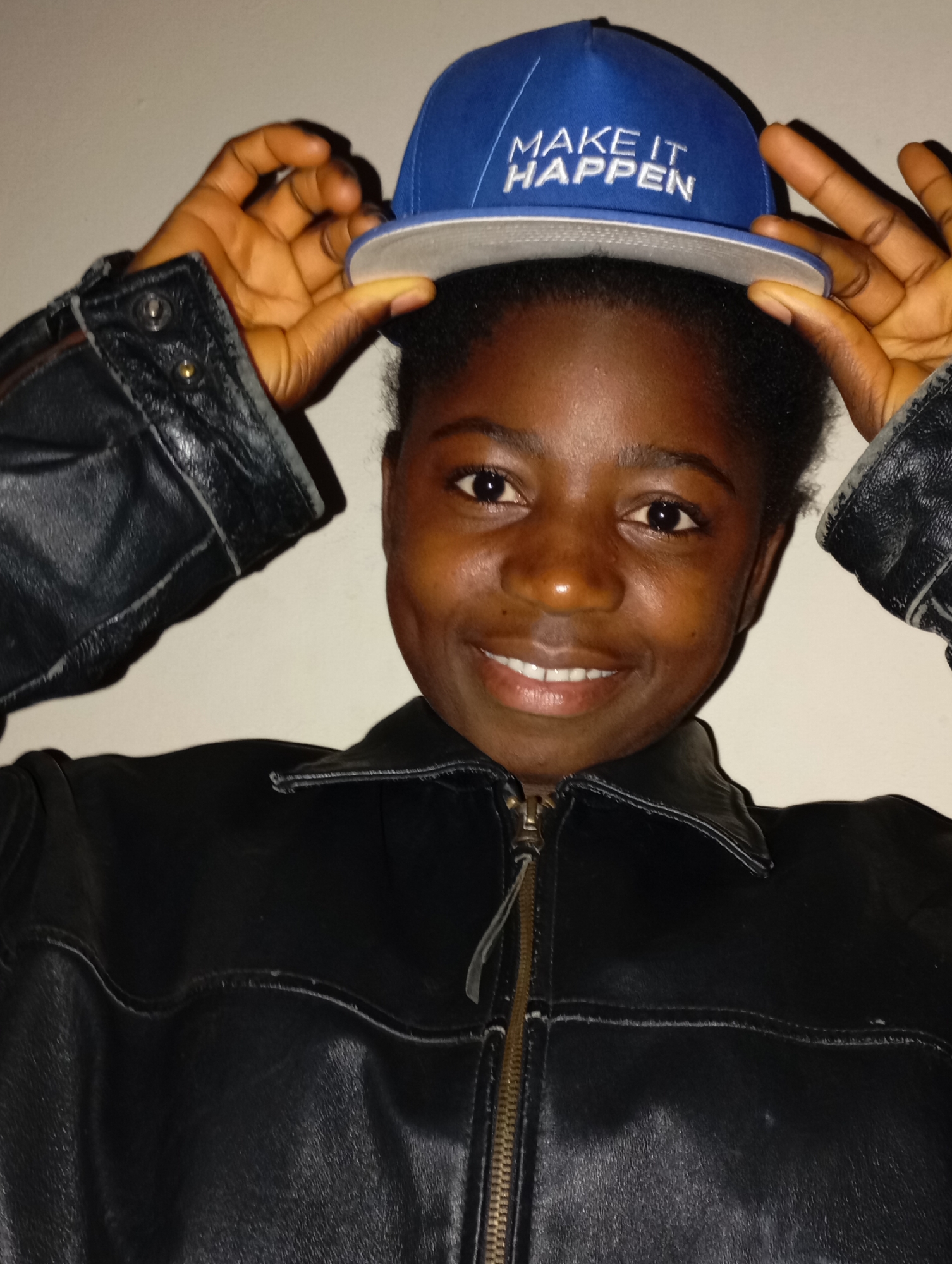 A dark-skinned woman is wearing a black leather jacket and holding a blue baseball cap with white text "MAKE IT HAPPEN" slightly above her head. The background is a plain, light-colored wall. It is not possible to confirm the location of the city of Jos, Nigeria.