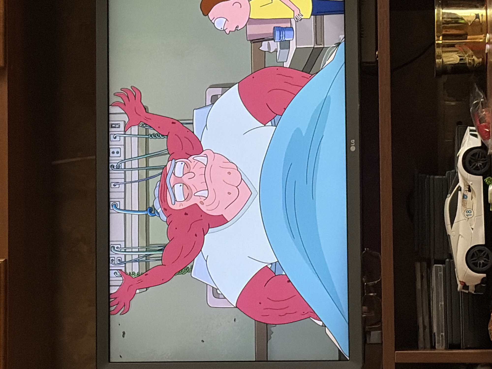 The image shows a television screen displaying a scene from the animated show Rick and Morty. In the foreground, a large, muscular, red-skinned alien with a pig-like snout and tusks is lying in a hospital bed, wearing a light blue v-neck shirt. Its arms are raised, and it appears to be either speaking or exerting effort. To the right, a small, boy-like character, identified as Morty Smith from the show, stands and looks towards the alien with a surprised expression. The background on the screen depicts a hospital room with medical equipment. Below the television, a shelf holds several DVD cases, a white toy sports car, a gold cylindrical container, and other miscellaneous items. The scene is set indoors, likely in a living room or entertainment area.