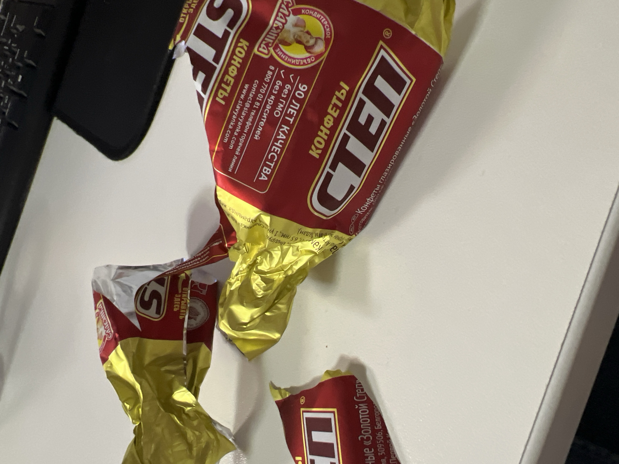 The image displays several crumpled wrappers from "СТЕП" (STEP) glazed candies by the Russian confectionery brand "Славянка" (Slavyanka). The wrappers, predominantly red and golden-yellow, are scattered on a plain, light-colored surface, likely a desk or table, with a dark, indistinct object visible in the upper left corner.
The main wrapper clearly shows "СТЕП КОНФЕТЫ" (STEP CANDIES) and specifies them as "Конфеты глазированные «Золотой Степ»" (Glazed candies "Golden Step"). The "Славянка" logo, featuring peanuts, is prominent, accompanied by "КОНДИТЕРСКОЕ ОБЪЕДИНЕНИЕ" (Confectionery Association). Text on the wrapper highlights "90 ЛЕТ КАЧЕСТВА" (90 YEARS OF QUALITY), "без ГМО" (GMO-free), and "без красителей" (without dyes). Contact information, including a phone number "8 800 770 01 81" and website "www.slavyanka.com", is also visible. Another wrapper fragment displays "ОТКРЫВАТЬ ЗДЕСЬ" (OPEN HERE).
The scene suggests that multiple candies have been consumed, leaving behind their empty packaging. The setting is an indoor environment, likely an office or home in Khimki, Russia, consistent with the Russian language on the packaging. No people are visible, and no specific time of day or weather can be discerned. The image captures the casual aftermath of enjoying these sweets.