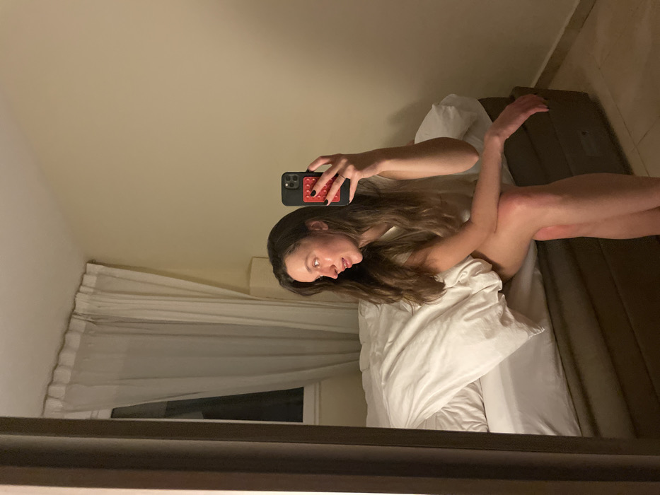 A woman lies on a bed covered with white sheets and pillows. She is taking a selfie with a phone. The background is a plain wall with a window partially covered by white curtains. The room appears to be lit by soft, indirect lighting. The image is taken from the perspective of a mirror reflecting the scene.