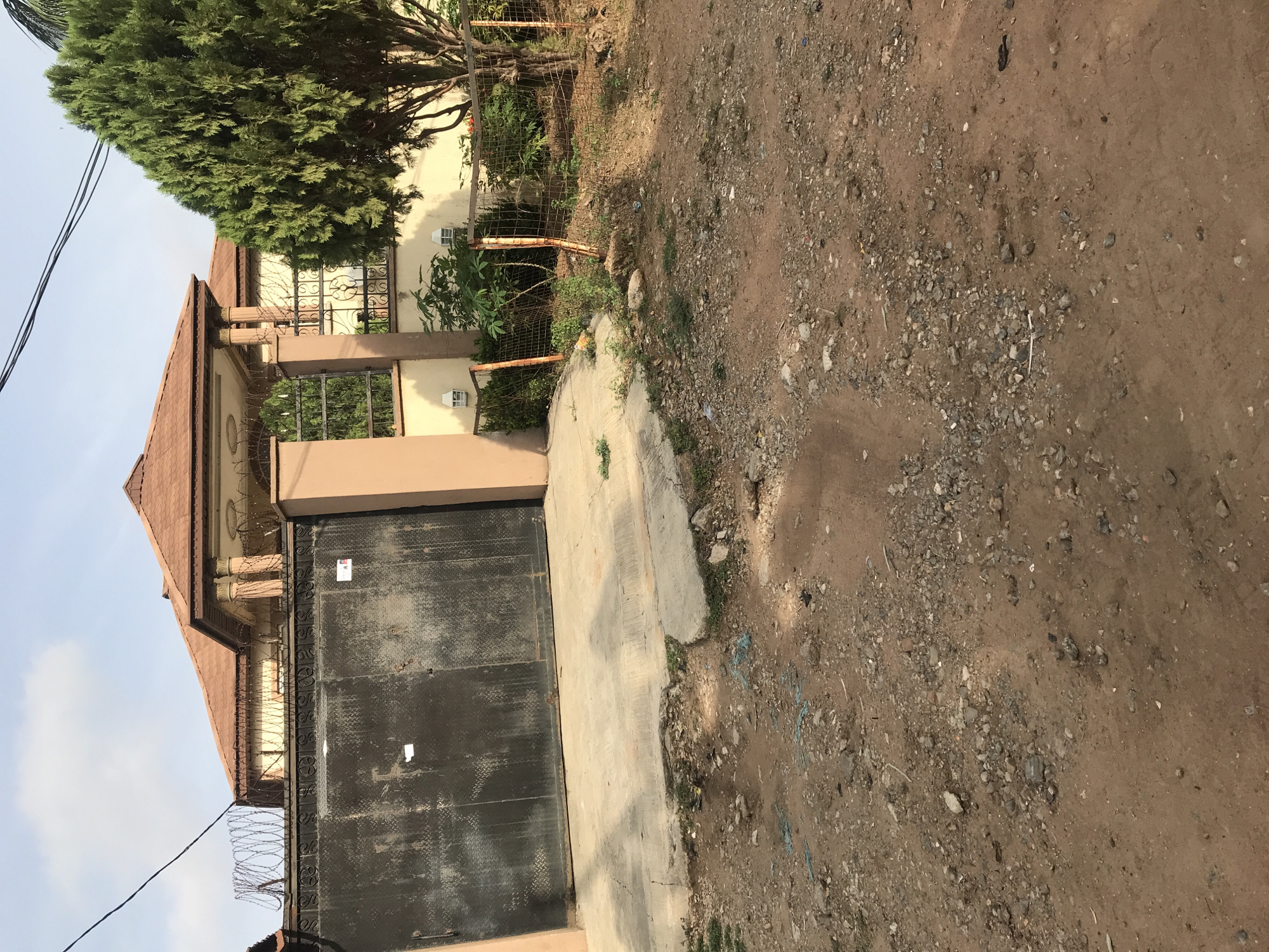 The image depicts the exterior of a fenced residential property in Oko Erin, Nigeria. The property features a large, black metal gate with visible wear and tear. A paved concrete pathway leads from the gate to the street. Above the gate, the building has a brown, tiled roof and tan-colored walls. Barbed wire is visible atop the gate and fence. A mature evergreen tree is to the right of the building. The street is unpaved, consisting of reddish-brown dirt and gravel. The sky is visible above the building, indicating daylight. No people are visible in the frame.
