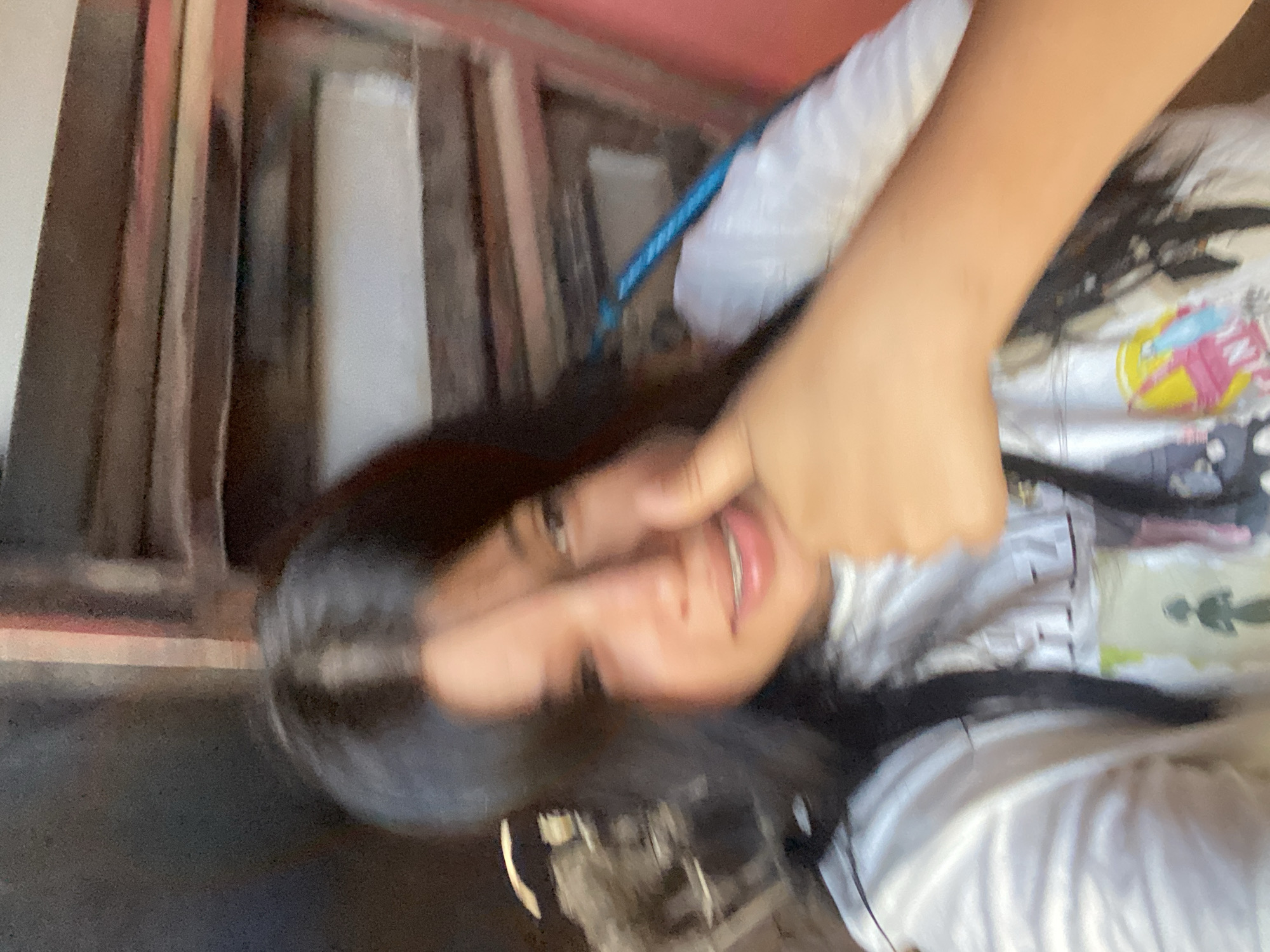 A person with long dark hair, fair skin, and braces on their teeth is positioned in front of what appears to be a window or door frame with wooden slats. The person is holding their hand near their face, possibly obscuring part of it. The background suggests an interior space. There is insufficient information to determine if the location is Juan Griego, Venezuela.