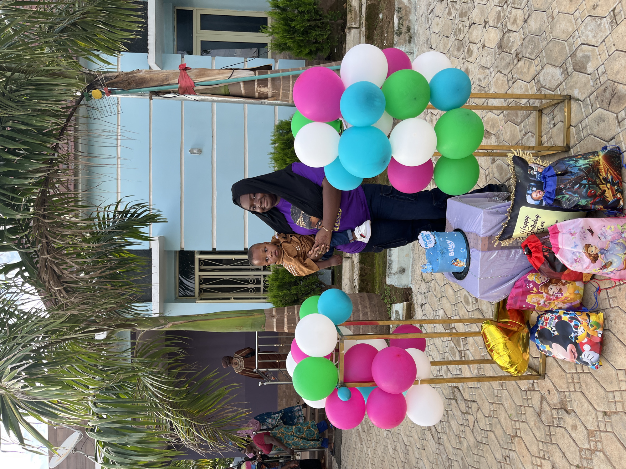 An outdoor birthday celebration is depicted in Jos, Nigeria, featuring an adult female holding an infant. The scene is set on an interlocking paved surface in front of a light blue painted building with white trim, featuring a barred door and windows. **People:** 1. **Central Female:** An adult female with dark skin, wearing a black head covering (hijab or scarf), a purple t-shirt with a partially visible "BLESSED" graphic, and dark trousers. She is smiling directly at the camera while holding an infant. Faint henna designs are visible on her left hand. 2. **Infant:** A male infant, appearing to be approximately one year old (supported by the cake topper "FAHEEM @ ONE"), with dark skin, wearing a light brown long-sleeved shirt and white sneakers. He is being held securely by the central female and is looking towards the right side of the frame with a neutral expression. 3. **Background Figures (Left):** Two adult females are visible on the left side. One wears a red top and a patterned blue-green skirt, walking left. The other, partially obscured, wears a head covering and a patterned blue dress, also walking left. Other indistinct figures are visible further in the background. 4. **Background Figure (Center-Left):** One individual, dressed in a brown traditional garment (possibly an agbada), stands on steps near a building entrance, facing away from the camera. **Objects and Decorations:** 1. **Balloons:** Numerous latex balloons in white, pink, green, and blue are arranged on two gold-colored metal stands positioned on either side of the central figures. 2. **Cake:** A two-tier round blue birthday cake, decorated with images of cartoon characters (Bluey and Paw Patrol), is situated on a black cake stand. A blue cake topper reads "FAHEEM @ ONE". The cake is placed on a table draped with purple fabric. 3. **Gifts/Favors:** Several themed bags for children, featuring characters such as Mickey Mouse, Disney Princesses, and Spider-Man, are arranged on the ground in front of the cake table. A gold foil balloon and a decorative cushion or pillow inscribed with "Happy Birthday Son" and a photograph of a child are also among the items. **Environment:** The event takes place outdoors, on a geometrically patterned paved area composed of brown and grey interlocking pavers. The background features a prominent light blue building with white architectural trim, equipped with a metal-barred security door and windows. Large palm trees with long, drooping fronds frame the scene,