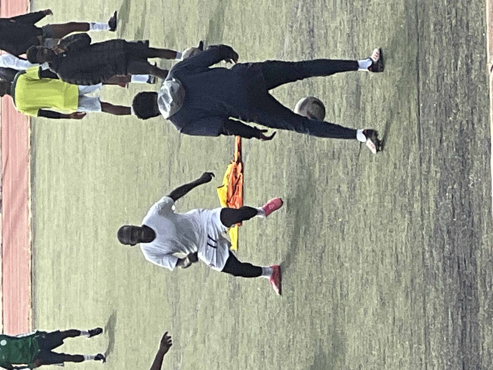 A group of young men, appearing to be soccer players, are gathered on a grassy field at the New Stadium in Nigeria. In the foreground, a player in a white t-shirt and shorts, with pink soccer cleats, is actively engaged with a soccer ball, his body positioned in a defensive stance. To his right, another player, also in dark athletic wear and soccer cleats, stands with a soccer ball near his feet, seemingly observing or preparing for a pass. In the background, other players in various athletic attire, including a green jersey, a yellow t-shirt, and dark jackets, are visible, some standing and others in motion. A pile of bright orange and yellow sports equipment is situated on the field between the two foreground players. The overall environment suggests a training session or a casual game taking place on a sunny day.