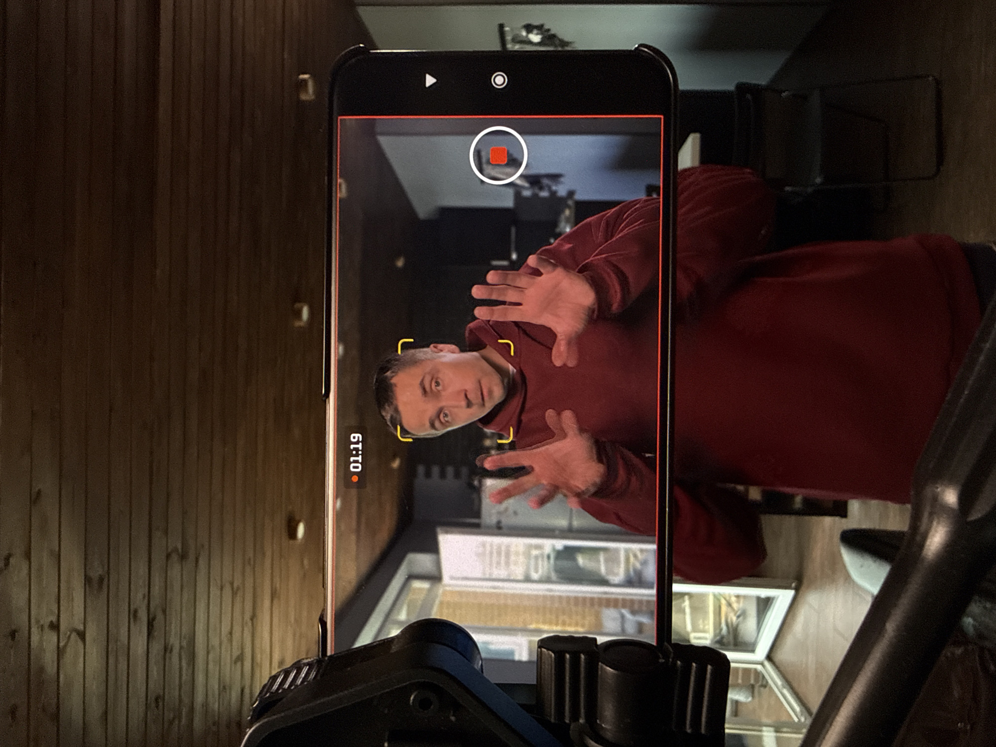 A smartphone, presumably mounted on a tripod, is recording a video. The video being recorded shows a man wearing a red hooded sweatshirt, his hands raised towards the camera. The background appears to be an interior scene, possibly a kitchen or dining area with a window visible behind him. The recording interface on the phone displays a timer reading "01:19" and a recording indicator at the top of the screen. The location is not visible from the current image.