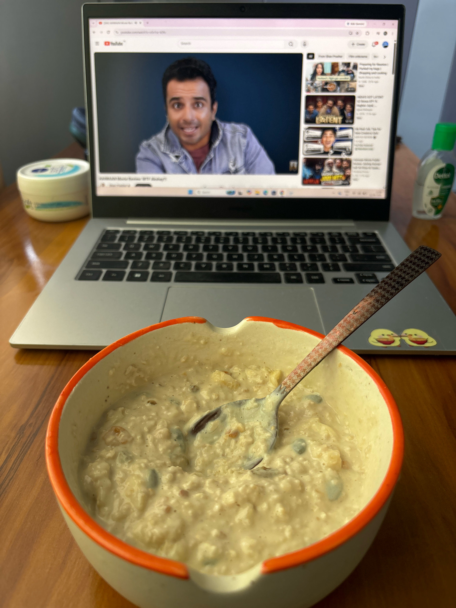 A medium shot captures a laptop screen displaying a YouTube video of a man in a denim jacket with a maroon t-shirt, looking surprised. Adjacent to the laptop is a bowl of oatmeal with a spoon, a tub of cream, and a bottle of hand sanitizer. The scene is set on a wooden table. The background suggests an indoor environment with a plain wall. The image lacks specific details to pinpoint its location within Bhatta, India.