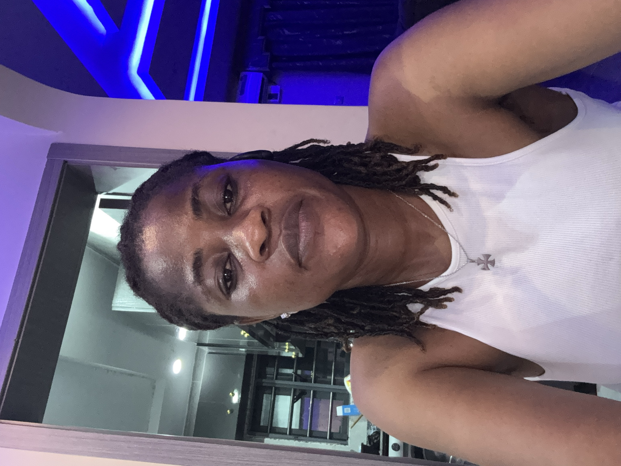 A medium shot selfie shows a Black woman with dark, braided hair and dark eyes looking directly at the camera. She is wearing a white, ribbed tank top and a silver necklace with a cross pendant. The background is dimly lit, with a window with a security grate visible on the left and blue LED lights on the right. The overall impression is that the photograph was taken indoors, likely in a residential setting. The location is not explicitly stated, but based on the presence of what appears to be a window with a security grate and the general aesthetic, it is plausible that this image was taken in the city of Ajara, Nigeria.