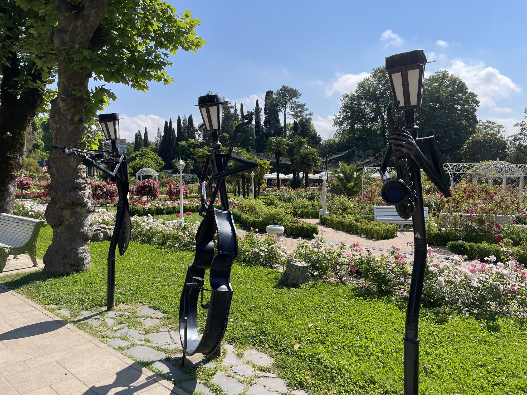 In an outdoor park setting on a sunny day, three tall, slender black metal sculptures stand prominently in the foreground. Each sculpture is designed to resemble a stylized human figure holding a musical instrument and has a black lantern on top. The sculpture on the left appears to be holding a cello. The middle sculpture is also holding a cello. The sculpture on the right is holding a saxophone.
Behind the sculptures, a vibrant garden is in bloom, featuring numerous rose bushes with pink and white flowers, as well as lush green hedges. A white park bench is visible in the middle ground. The background consists of a dense array of trees, including both deciduous and coniferous varieties, and a clear blue sky with scattered white clouds. The ground in the foreground is a paved walkway with a pathway of grey stones leading into the grassy area with the sculptures. The overall scene suggests a well-maintained public park or botanical garden.