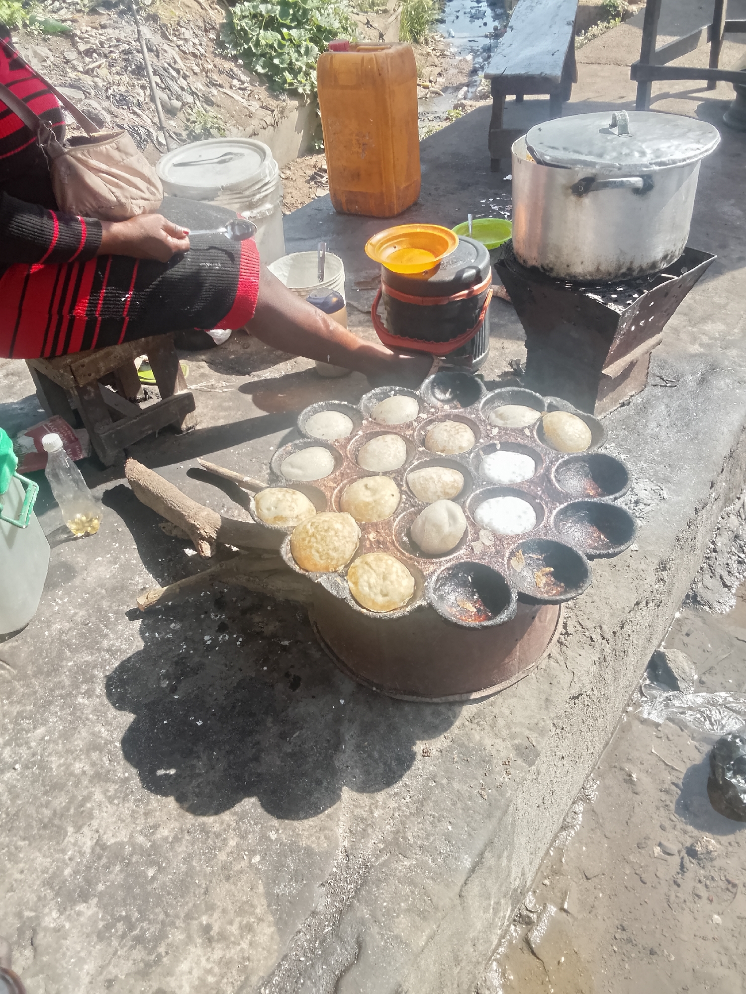 The image depicts a street food vending scene in an unknown city. A person seated on a low wooden stool is making small, round cakes in a cast iron pan with multiple cup-like depressions. Some cakes are cooked golden brown, while others are still in batter form. Next to the pan is a plastic bottle, a can, and a metal pot sitting on a small coal-burning stove. Other items include a water container and a jug, with benches in the background. The location is outdoors, on a concrete surface adjacent to a muddy area.