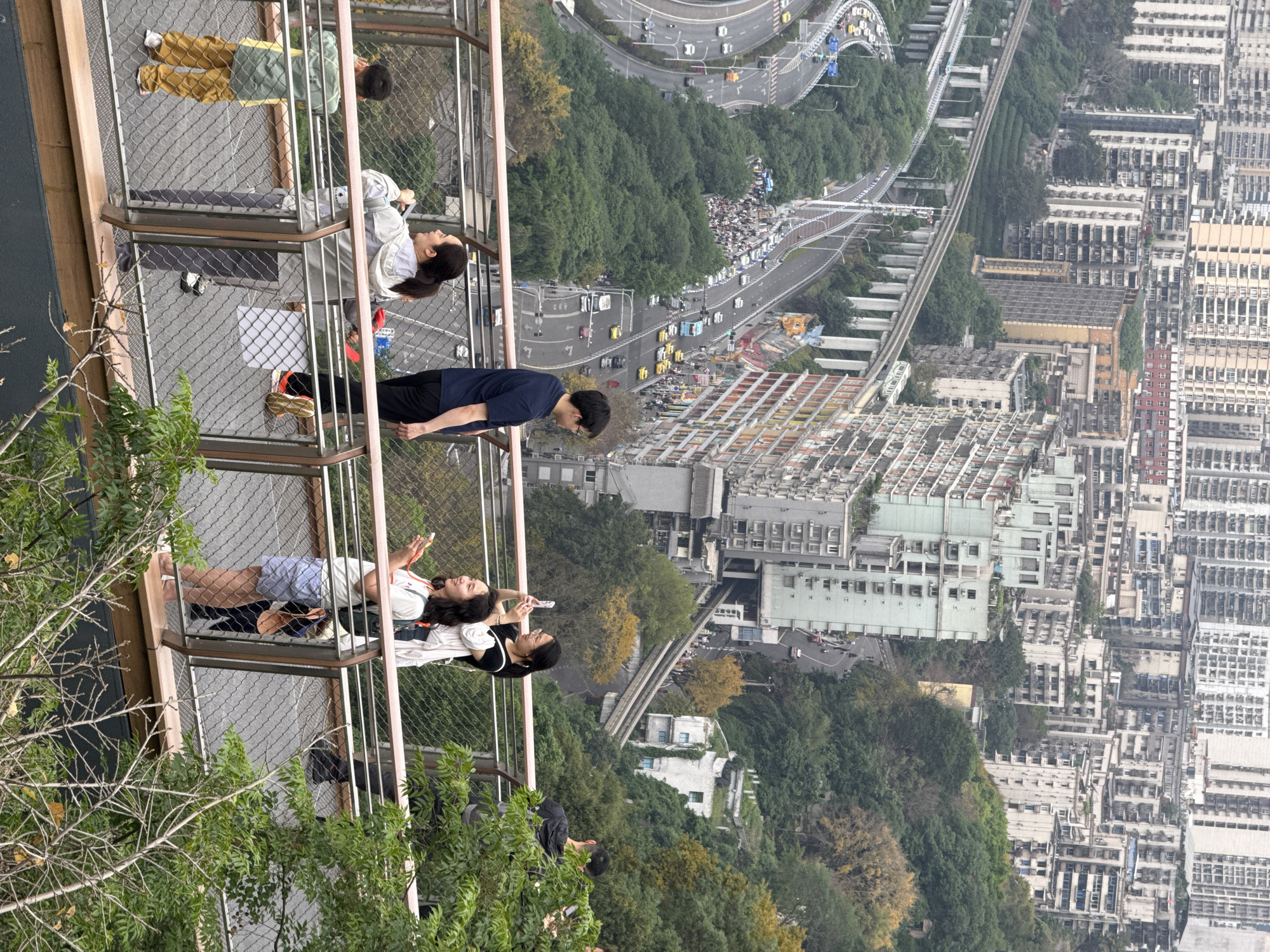 The image shows a group of people standing on multiple levels of a lookout platform in Yuzhong District, Chongqing, China. The platform overlooks a cityscape with a road, elevated railway, trees, and buildings. On the platform, people are seen standing, leaning, and lying down, with some using their phones. The foreground includes parts of the platform's structure and some tree branches.