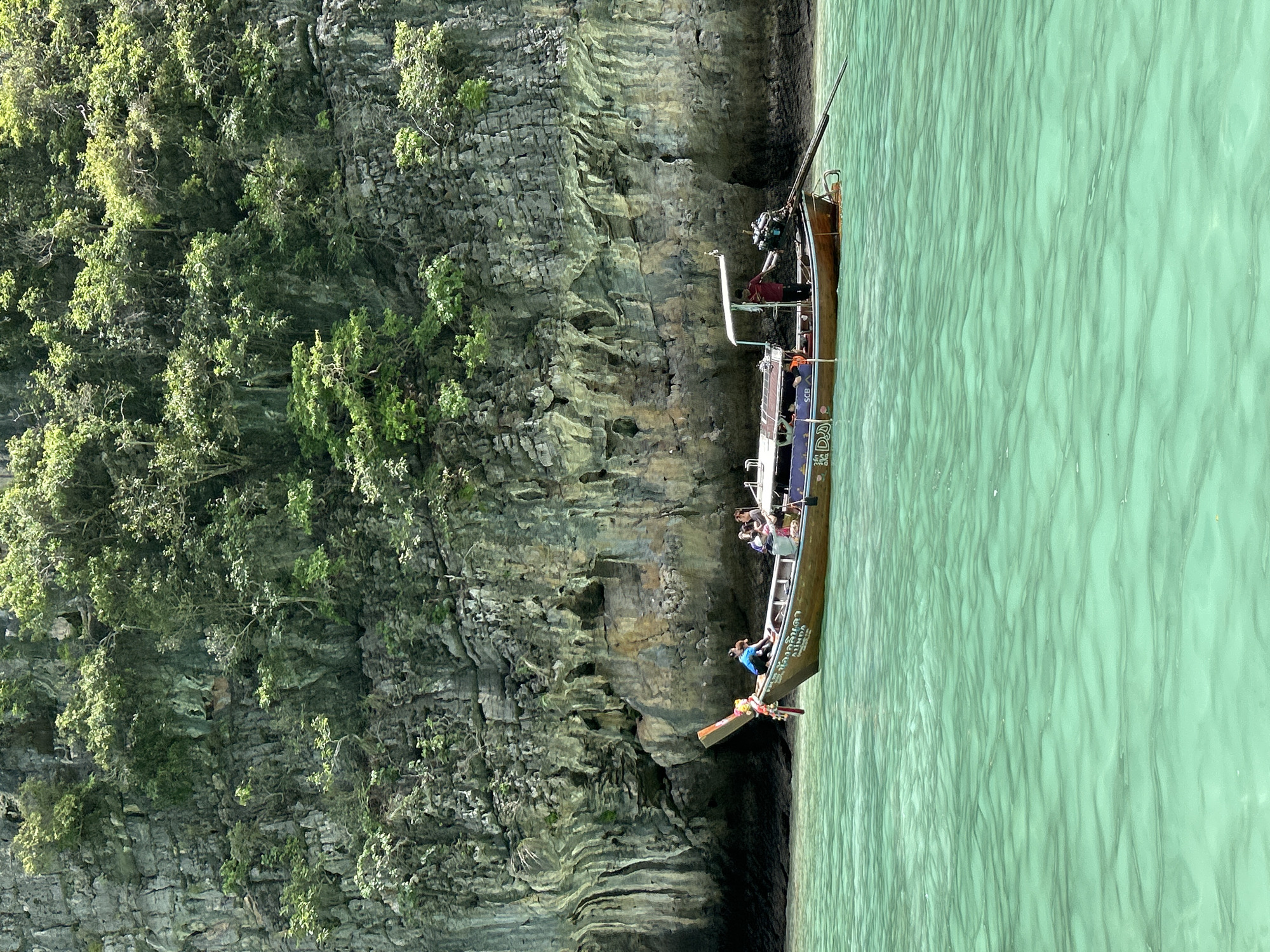 A longtail boat, a traditional Thai vessel, navigates the clear, turquoise waters near a limestone cliff face in Ao Nang, Thailand. The cliff is covered in lush green vegetation, with trees and shrubs growing out of its rocky surface. The boat has several people on board, including a person steering at the back, and others seated or standing. The boat is wooden, painted in brown and blue, and has an outboard motor at the stern. The water is calm with gentle ripples. The scene depicts a typical tourist excursion in the region, known for its stunning karst landscapes and clear waters.