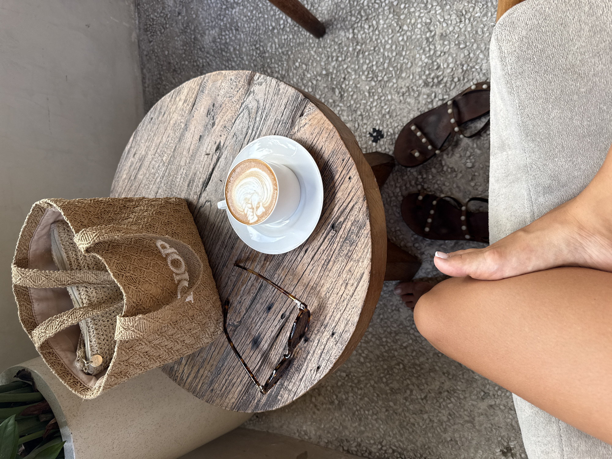 Overhead shot from an elevated viewpoint, focusing on a small round wooden table situated in an outdoor setting. The table is positioned on a light grey concrete floor. On the table, there is a white cup of latte art on a white saucer, a pair of brown tortoiseshell sunglasses, and a tan woven tote bag with "DIOR" written in white. Near the table, there is a pair of brown studded sandals. The leg and bare foot of a person seated are also visible to the right of the table, appearing tan in skin tone. The scene suggests a cafe or outdoor space in North Kuta, Indonesia.