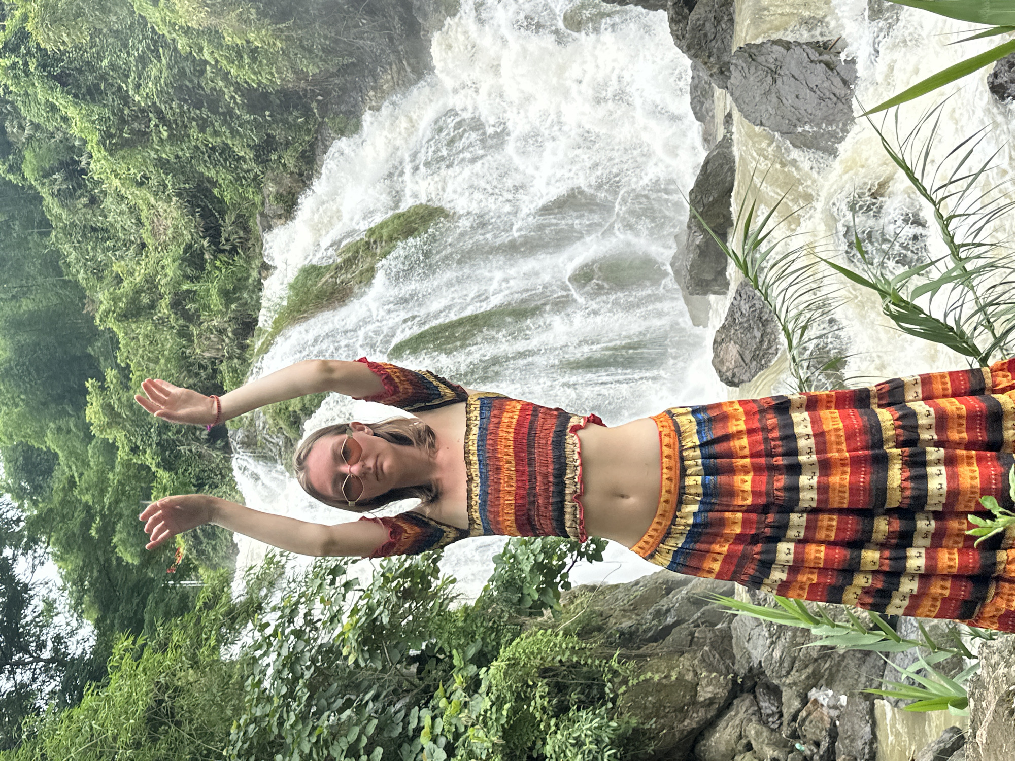 A young woman stands in front of a cascading waterfall in Bản Phúc, Vietnam, with her arms raised and hands open. She is wearing a colorful, patterned crop top and a matching tiered skirt. The woman has blonde hair and is wearing sunglasses. The waterfall is surrounded by lush green vegetation and large rocks. The water appears turbulent and a light brown color.
