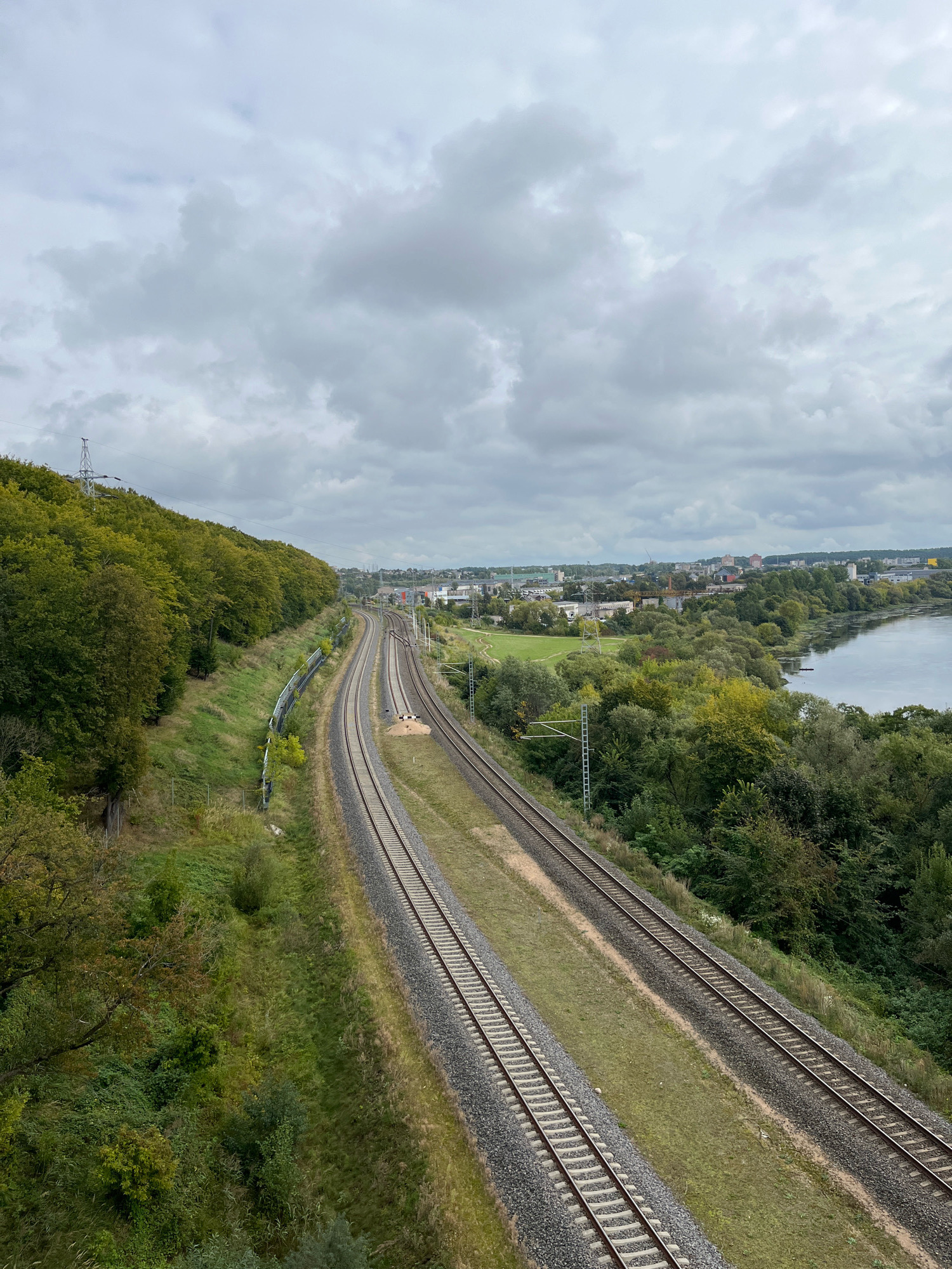 The image shows a landscape view of the city of Kaunas, Lithuania, featuring multiple railway tracks running through a verdant environment. On the left side of the frame, a tree-covered hill slopes upwards, with dense green foliage characteristic of a temperate climate, possibly in autumn given the varied green hues. A power line pylon is visible on the crest of the hill.
The railway tracks, appearing well-maintained, are a prominent feature, with two sets of parallel tracks diverging and curving into the distance. They are laid on ballast, with grassy verges on either side.
To the right, the landscape opens up to reveal a body of water, likely a river or a lake, bordered by a mix of trees and shrubs, some of which show hints of autumnal color. Beyond the water, a cityscape is visible, with various buildings, industrial structures, and what appear to be residential areas. The sky is overcast with heavy, grey clouds, suggesting a subdued lighting condition. The overall scene captures a blend of natural landscape and urban infrastructure, typical of areas where nature and human development coexist.