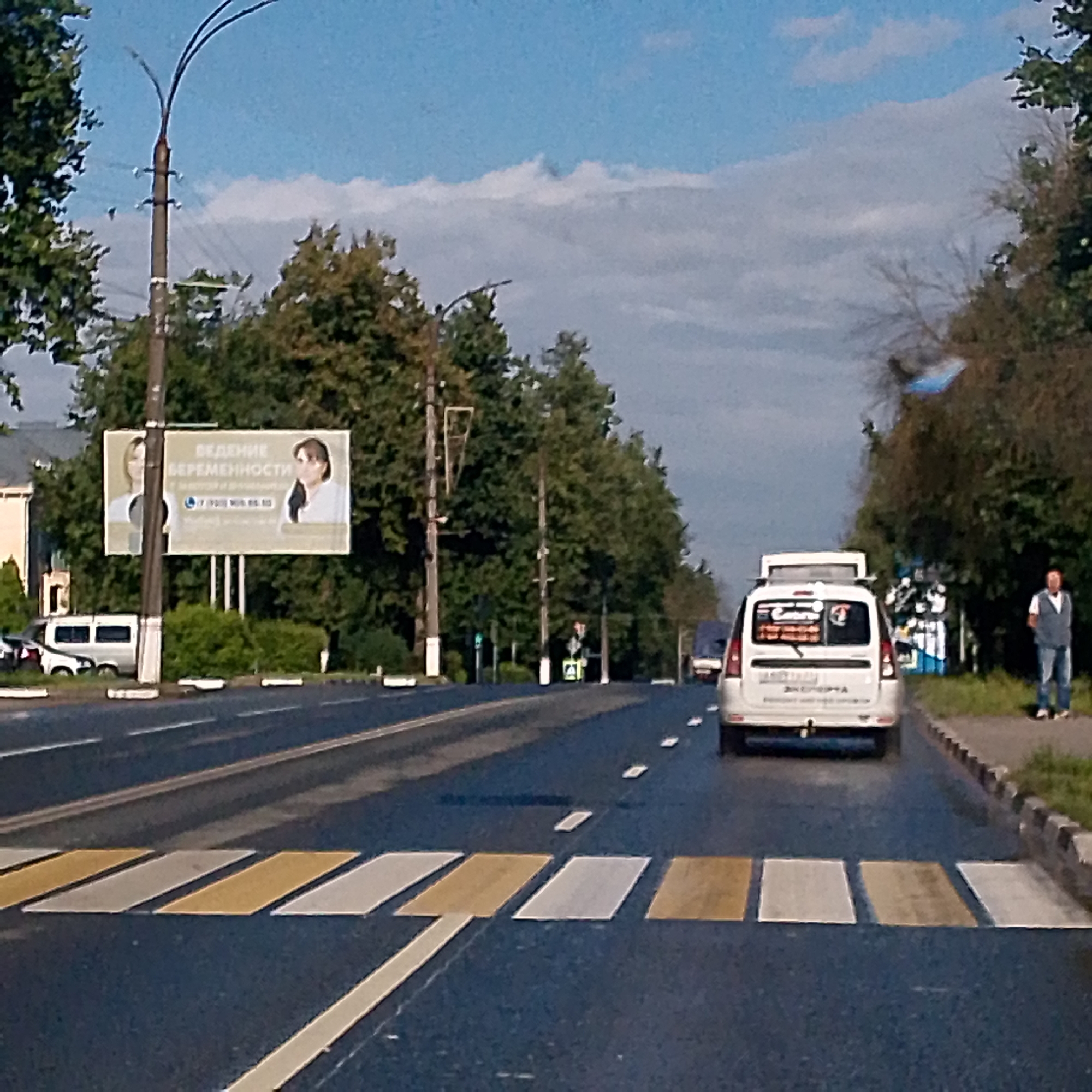 The image depicts a street scene in the city of Vyazniki, Russia, likely during the daytime. In the foreground, a pedestrian crossing with alternating yellow and white stripes is visible on a dark asphalt road. Cars are present on the road; a white minivan is moving away from the camera, and a white van is stopped in the distance. On the right side of the road, a man in a light blue vest and jeans stands on the sidewalk, which is bordered by grass and a curb. Further down the street, trees line the road, and a billboard advertising "Bearing Pregnancy" is visible. A lamppost stands near the billboard. The sky above is a mix of blue with scattered white clouds.