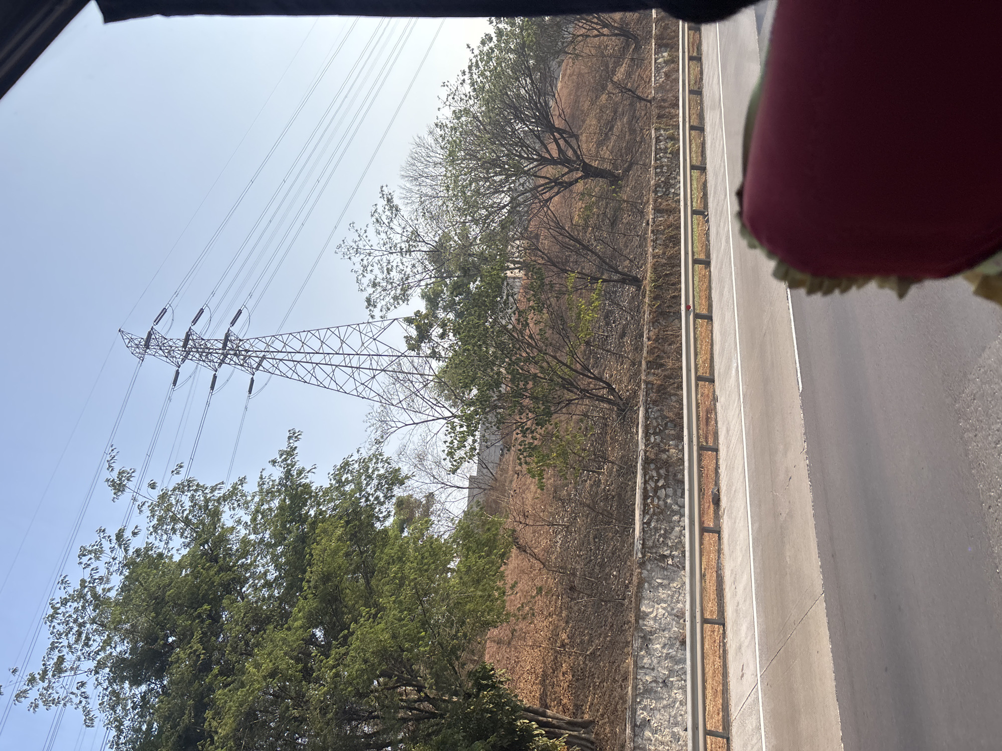 The image depicts a roadside view with a high-voltage electricity pylon and power lines dominating the sky. The pylon, a lattice steel structure, is positioned on a dry, sloping hillside covered with sparse vegetation and fallen leaves. A retaining wall made of rough-hewn stones runs along the base of the hill. In the foreground, a highway or major road is visible, with a metal guardrail separating it from the embankment. The sky is clear and blue, suggesting daytime. The image appears to have been taken from a moving vehicle, as indicated by the blurred foreground and the dark frame on the right side of the image, likely from a window or pillar. The scene is devoid of people or any discernible activity. Based on the presence of such infrastructure, the image could be located in an industrial or semi-urban area of Karawang, Indonesia, a region known for its industrial estates and power transmission networks.