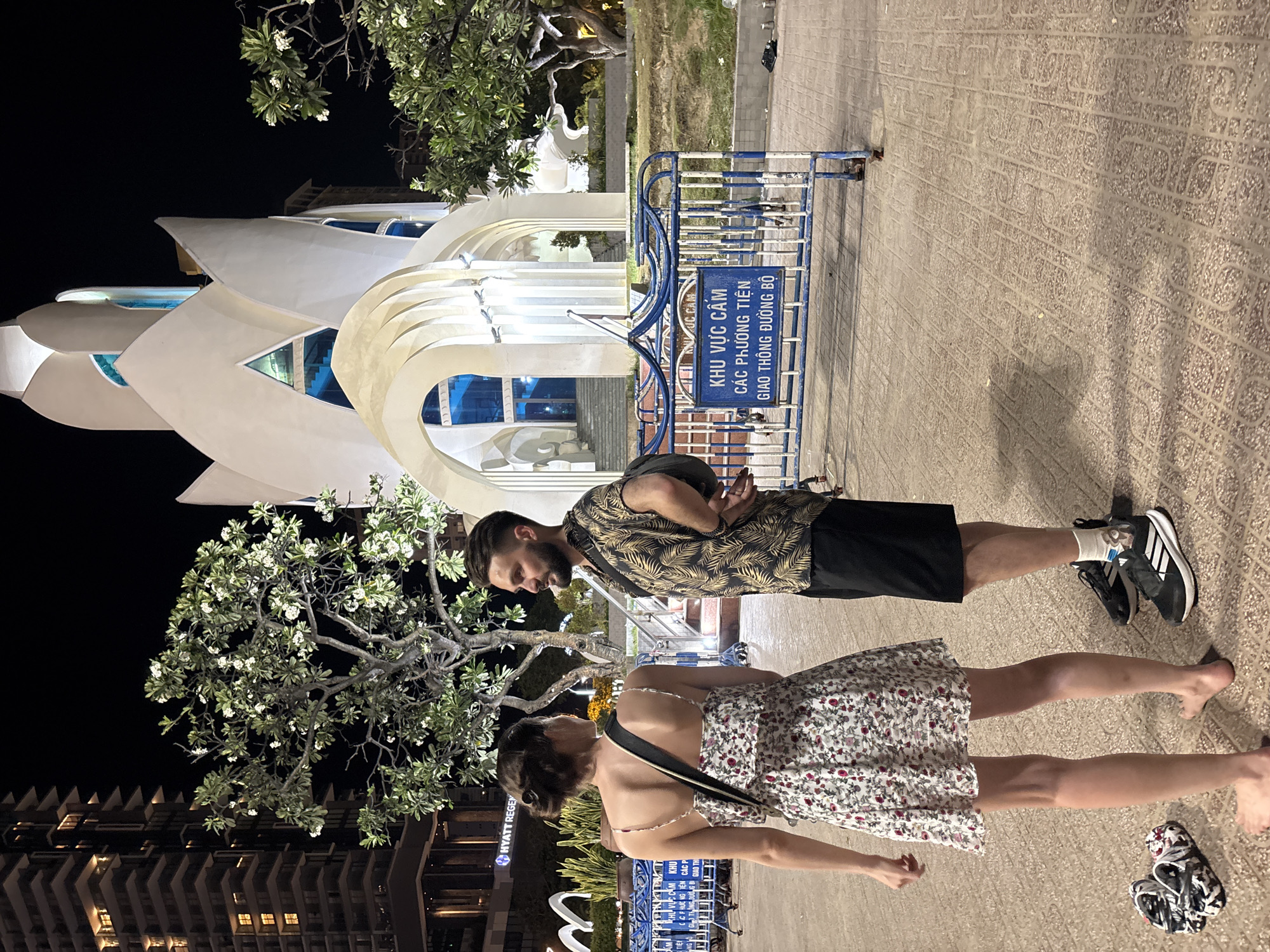 In the city of Nha Trang, Ward, Vietnam, at night, a man and a woman stand facing each other on a paved plaza. The woman is on the left, wearing a floral print dress and a crossbody bag. Her hair is pulled back in a bun, and she is barefoot. The man is on the right, wearing a short-sleeved, patterned shirt and black shorts. He has a beard and is wearing sneakers.
Behind them, a distinctive white lotus-shaped building with blue-tinted windows is illuminated. To the left of the building, a tall, modern building with many illuminated windows stands against the dark night sky. In the foreground, a blue and white metal barrier with a sign in Vietnamese is present. Trees with green leaves and white flowers are visible throughout the scene, casting shadows on the plaza. The ground is paved with textured tiles. A pair of sandals is placed on the ground near the woman's feet.