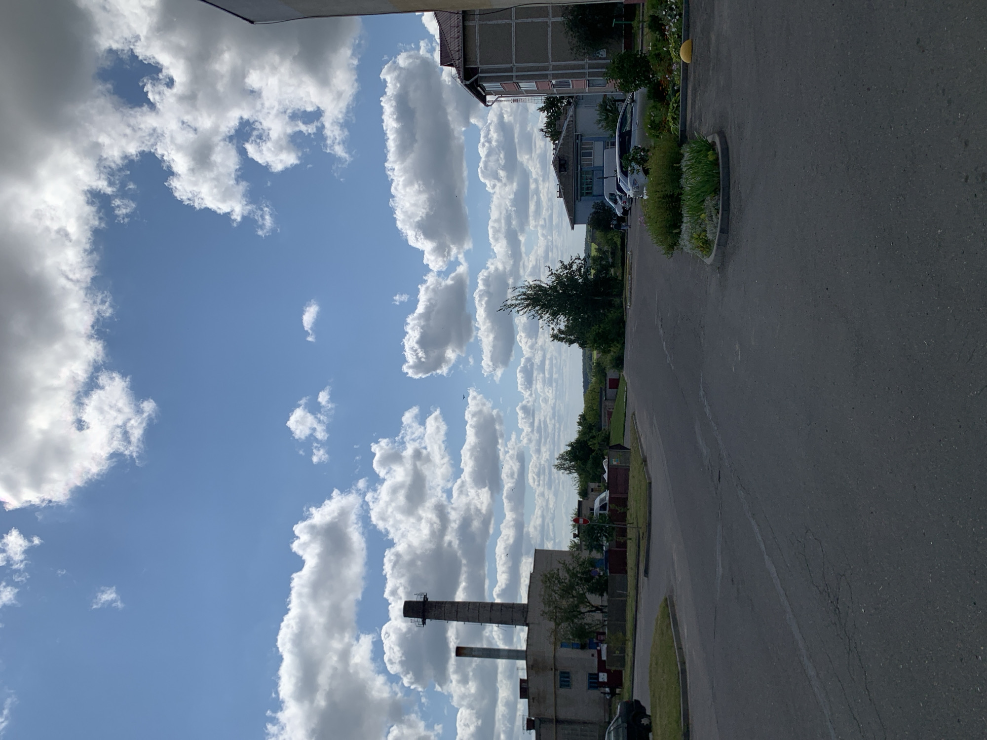 The image depicts an overcast day in Kapyĺ, Belarus, showcasing a street scene with industrial and residential elements. In the foreground, a paved road with visible cracks and some patching suggests typical urban wear. To the left, a black car is parked, partially obscuring a grassy area and a low fence. Dominating the mid-ground on the left is a large industrial building with two tall smokestacks, one significantly taller than the other, constructed from aged brick. Adjacent to this building is a smaller structure, also appearing to be part of the industrial complex.
Further into the mid-ground, a road curves to the right, leading towards a cluster of residential buildings. One building, taller and more modern, displays balconies and a tiled roof. Across the street, a smaller, older building with a grey facade and a blue-trimmed window is visible, with a white van parked in front of it. Several trees are scattered throughout the scene, including a tall, slender tree directly behind the industrial building.
In the background, a soft, hazy landscape of rolling hills and greenery stretches out under a bright blue sky filled with voluminous white cumulus clouds. The overall impression is one of a functional, somewhat industrial area of the city on a bright but cloudy day. The presence of parked cars and the general upkeep of the residential areas suggest ongoing daily activity.