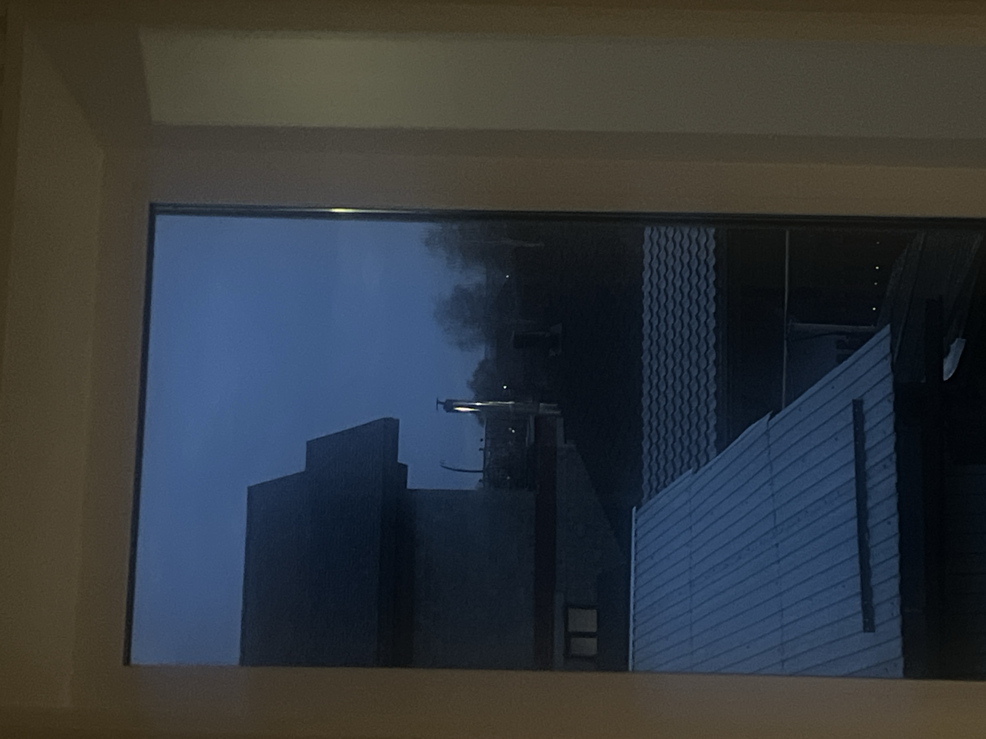 View from a window frame onto a dark, industrial scene in Votkinsk, Russia. Visible are buildings: one a stepped tower, another with a corrugated facade, and part of a vehicle on the right edge. Smoke plumes rise behind the structures. It is dusk or night.