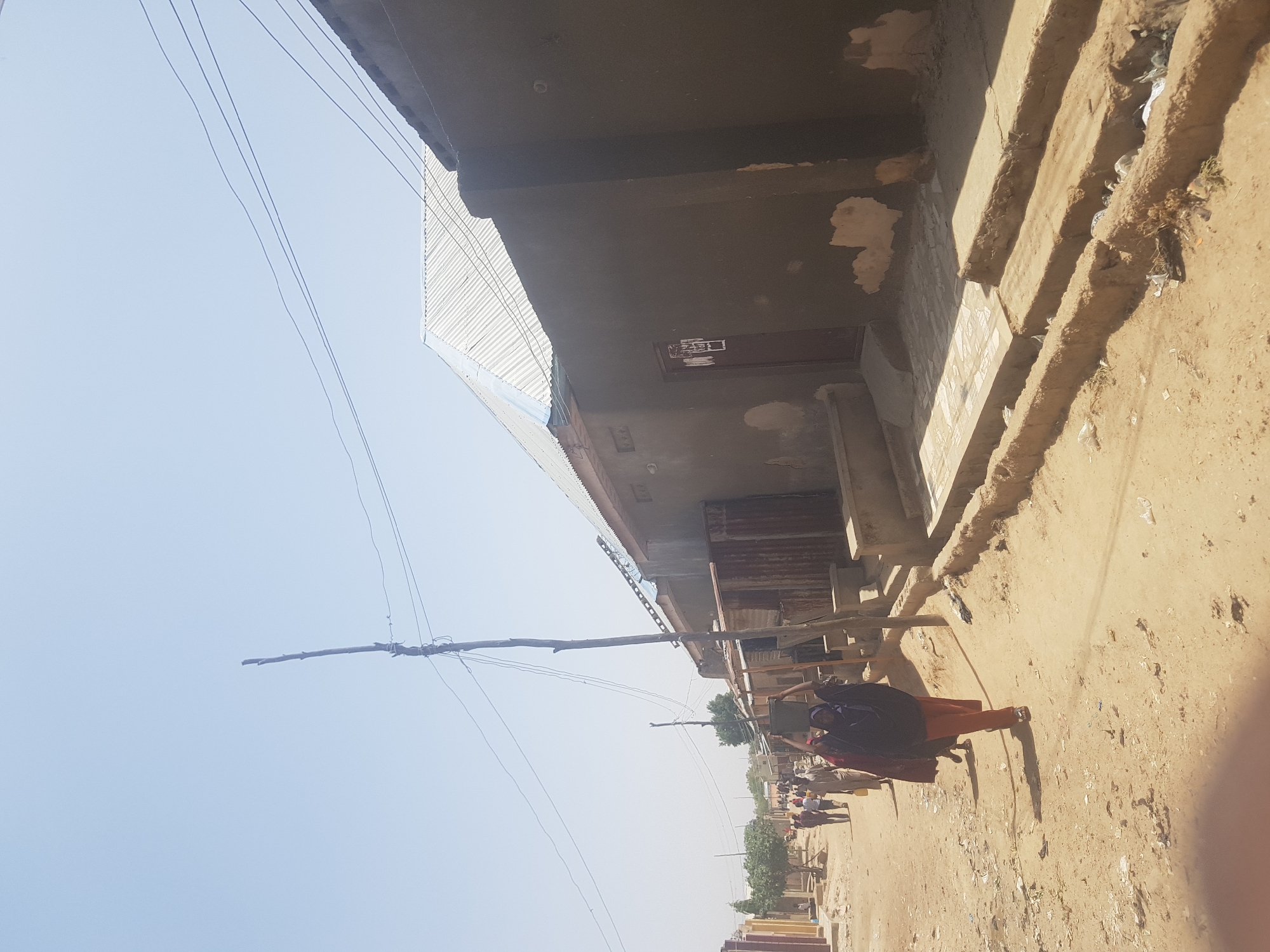 The image shows a street scene in Dutsinma B, Nigeria. The ground is a light tan, sandy dirt. A building with a gray façade and a corrugated metal roof is on the left. Power lines run along the top left. People are walking along the street. One person is wearing a dark purple or black hijab and an orange dress. Others are gathered further down the street. The sky is light blue.