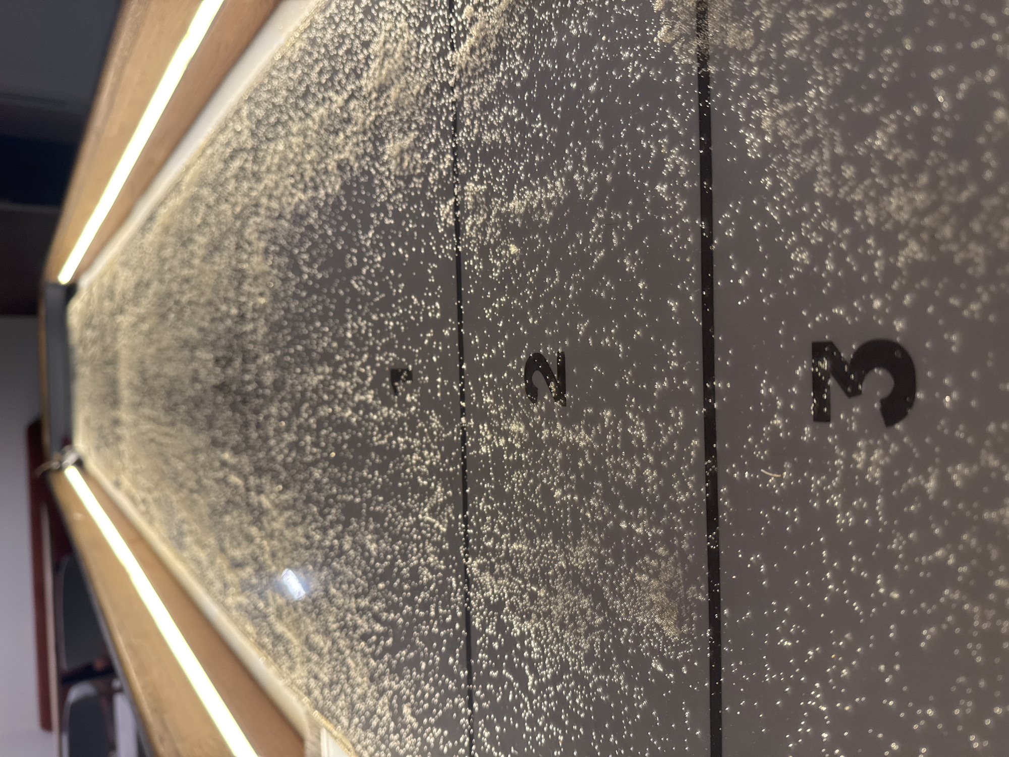 The image shows a long, rectangular light panel with the numbers 1, 2, and 3 visible on its surface. The panel is bordered by a wooden frame with embedded LED strip lighting. The surface of the panel is covered in what appears to be a fine, granular material. The numbers are dark and slightly offset from the central lines. The location and subject are not discernable from the image.