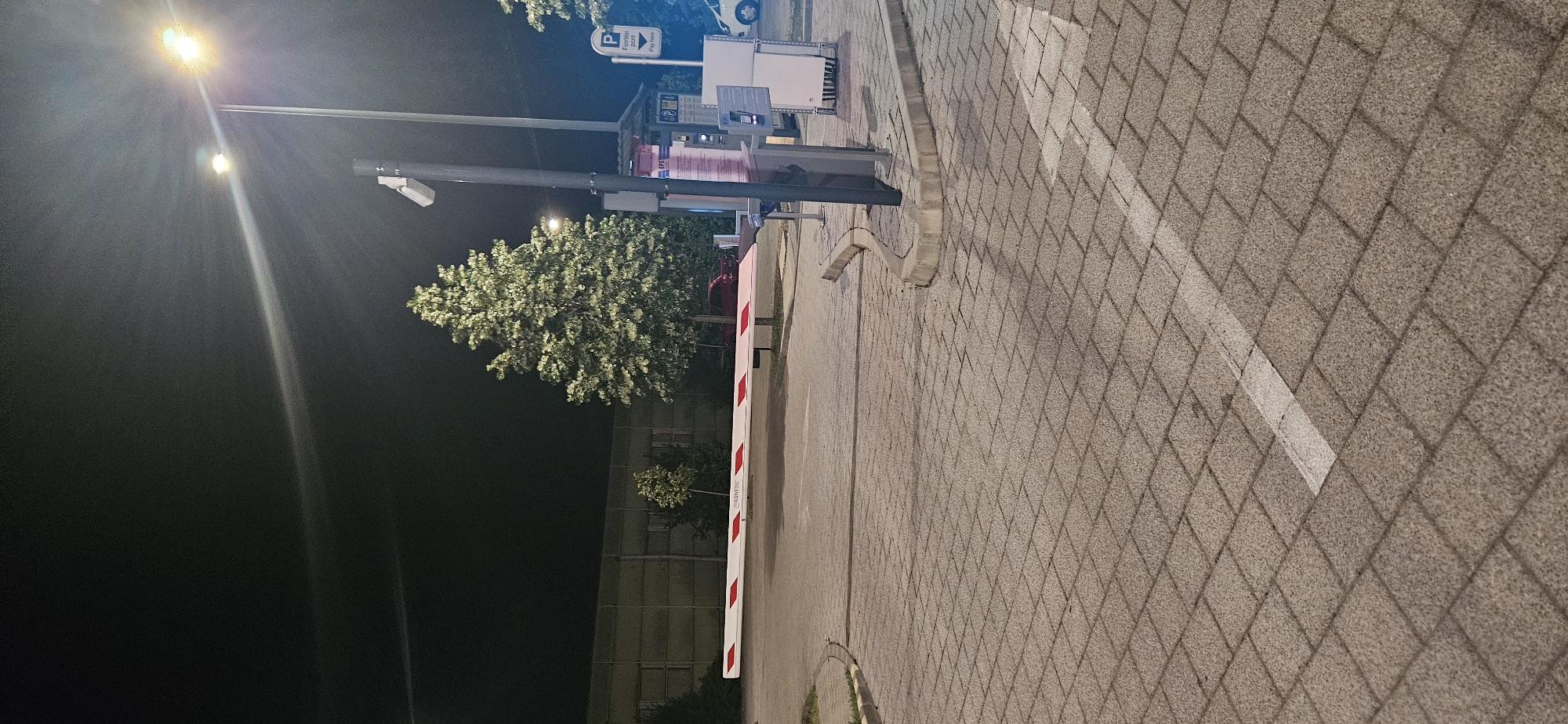 The image depicts a nighttime scene in a parking lot in Hévíz, Hungary. A white barrier arm with red diagonal stripes is lowered, blocking an entrance. To the right of the barrier, a payment station with a touchscreen and ticket dispenser is visible, along with a security camera mounted on a pole. A large, leafy tree is illuminated by a street light, casting shadows on the paver stone ground. In the background, a light-colored building with a corrugated metal facade is partially visible. A red car is parked behind the barrier. The sky is dark, with a bright street light creating a lens flare effect.
