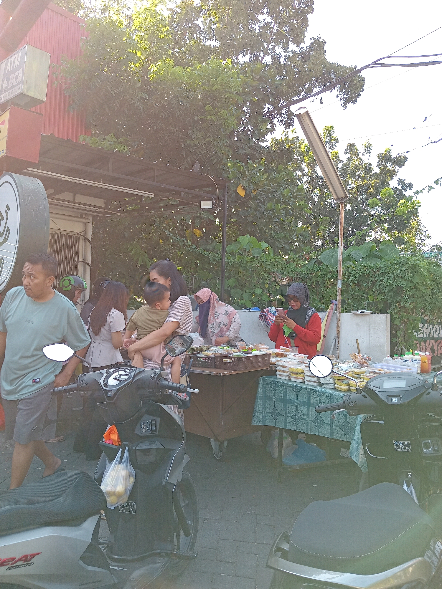 In Randuagung, Indonesia, a street scene unfolds with a vendor displaying food items on a table. The vendor, wearing a red shirt and a grey headscarf, is looking at her phone. Behind the table, various food containers are neatly arranged. To the left, a woman is holding a child, and other individuals are present, some appearing to be customers. Motorcycles are parked nearby, one prominently in the foreground. The environment suggests a public space, possibly a market or roadside stall, with buildings and lush greenery in the background. The time of day appears to be late afternoon or early evening, given the lighting conditions.