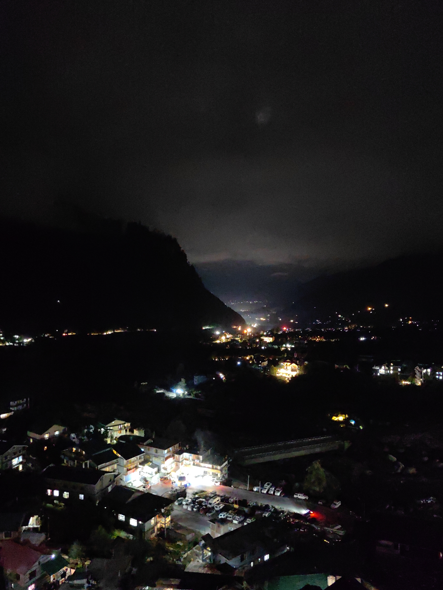 An aerial nighttime view captures a valley town illuminated by artificial lights. On the left, a dark mountain looms, its peak obscured by clouds. The town itself is nestled in the valley, with buildings and roads visible. A bridge spans across the valley in the middle ground. Numerous cars are parked in open lots and along roads. The overall scene suggests a residential and commercial area within a mountainous region. The location is identified as Kulang, India, implying a mountainous terrain and typical Indian architecture.