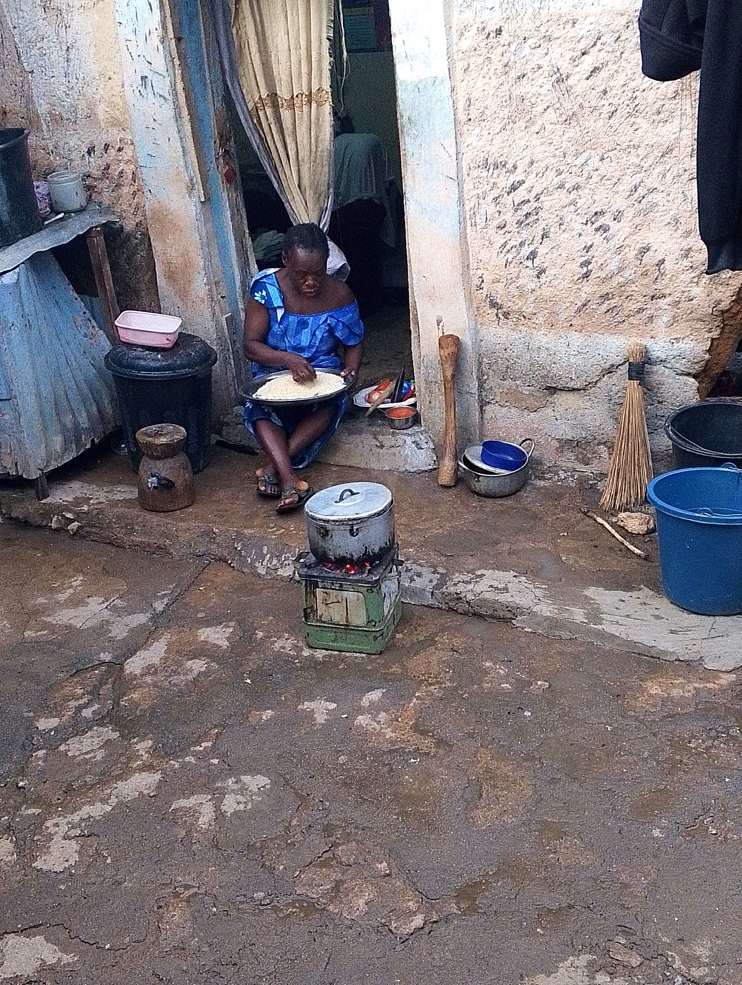The image depicts a woman sitting on a stoop in what appears to be Jos, Nigeria, preparing food. The woman, wearing a blue dress, is holding a plate of rice. A pot is cooking on a small stove in front of her. Various containers, kitchen implements, and a broom are scattered around the immediate area. The ground is a cracked, uneven concrete surface. The doorway behind the woman is partially obscured by a curtain. The building itself is constructed of a rough, textured material and shows signs of age and weathering.