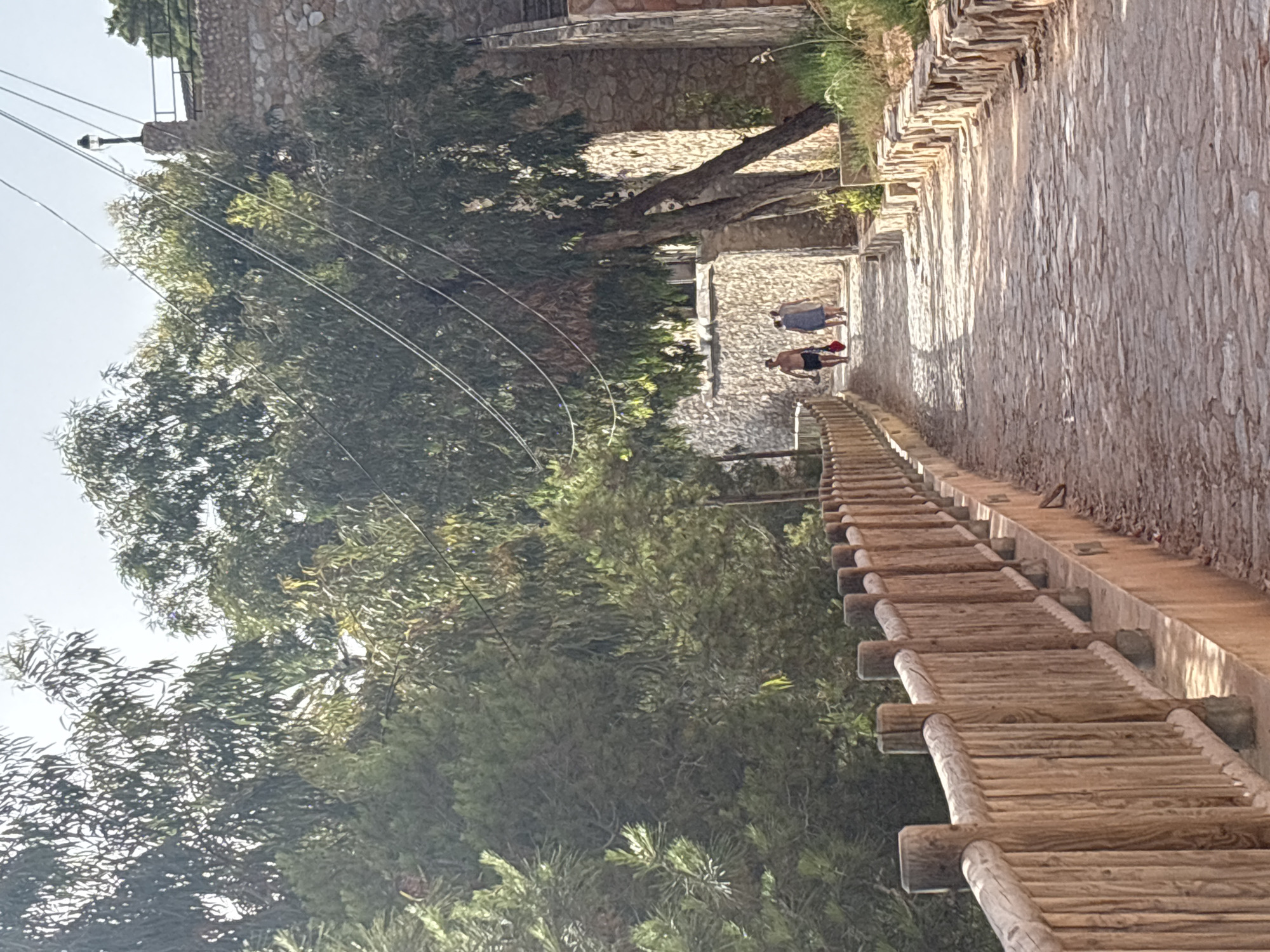 The image depicts a sun-drenched, cobblestone pathway in Urbanización Cabo Roig, Spain. The pathway is lined on the left by a rustic wooden fence and on the right by a stone wall topped with vegetation. Trees with dense green foliage overhang the pathway, creating dappled shade. In the distance, three individuals are walking away from the camera up the path. The person in front, a male, is wearing shorts and carrying a red bag. Behind him, two individuals, likely female, are dressed in light-colored clothing. A lamppost is visible on the right side of the image, atop a stone structure. The overall scene suggests a warm, outdoor environment, possibly a scenic route within the residential area.