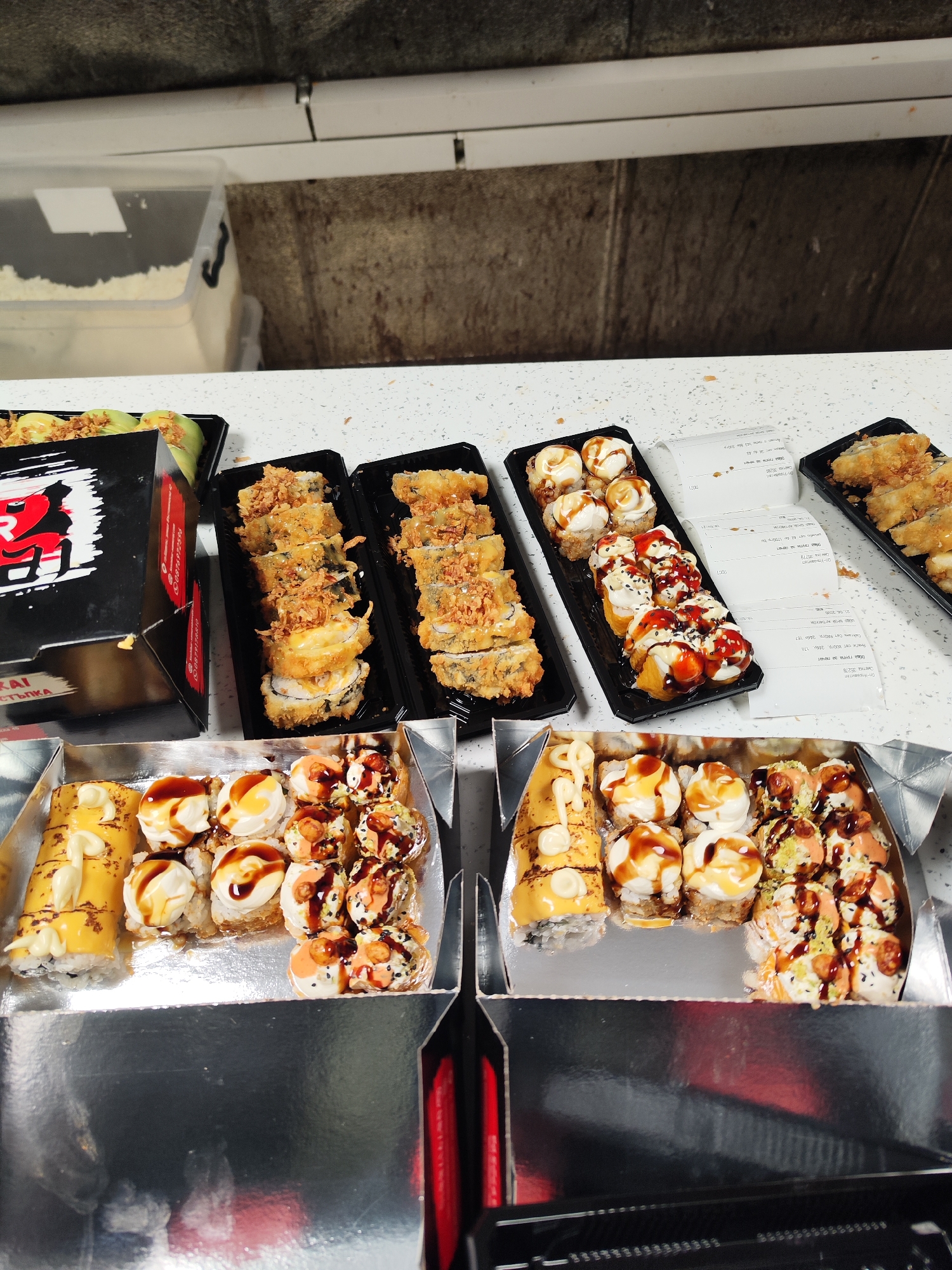 The image displays a variety of sushi rolls arranged on a white countertop, likely part of a large takeout or delivery order from a restaurant in Burgas, Bulgaria. The main subjects are numerous sushi trays and boxes filled with different types of sushi.
Two long black trays feature deep-fried or tempura-battered sushi rolls, generously topped with crispy fried onions. Another black tray holds rolls covered in white cream cheese or mayonnaise, drizzled with a dark sauce. Two open, metallic-lined cardboard boxes contain additional sushi selections: one box features a large, yellow-wrapped roll alongside rolls with cream cheese and dark sauce, while the other holds rolls with diverse toppings, sauces, and sesame seeds.
A partially visible black cardboard box on the left, branded with "RAI" and "СТЪПКА" (Step) in red, also shows a phone number like "0876972898". Scattered among the sushi trays are several crumpled paper receipts. Visible text on these receipts includes "Оп-Управител" (Manager), "Сметка 35278" (Bill 35278), "Сет PROгр. 2460 174" (Set PROgr. 2460 174), and the date "21.04.2026". Another receipt clearly states "Сметка 35280" (Bill 35280).
The background consists of a dark, textured wall, possibly concrete or a similar industrial surface, with a white horizontal pipe or conduit running across it. A transparent plastic container with white contents is visible in the upper left background. The lighting suggests an indoor setting. No people are visible in the scene. The presence of multiple receipts and varied sushi types implies a busy service or a substantial order being prepared or awaiting collection.
