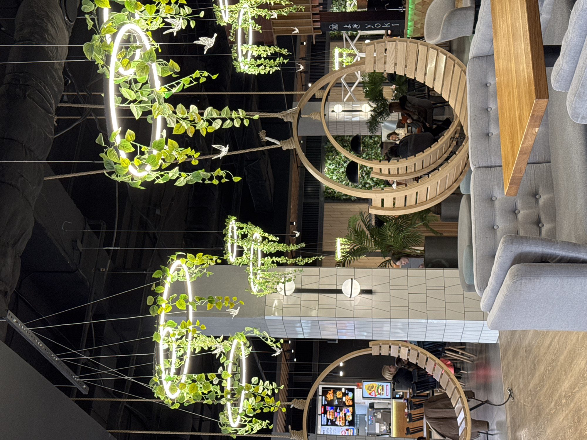 The image captures the interior of a modern and stylish food court or restaurant, likely located in Izhevsk, Russia.
The most striking visual elements are the overhead decorations: a series of circular, neon-lit rings heavily entwined with lush artificial green vines. Small, white bird-shaped ornaments are interspersed among the foliage, and these decorative elements are suspended from a dark, high ceiling by thick, knotted ropes, giving a contemporary, nature-inspired feel.
Seating arrangements are varied and modern. On one side, there are comfortable grey upholstered sofas and armchairs featuring button tufting, paired with a clean wooden table. On the opposite side, unique, curved wooden structures create semi-enclosed booth-style seating, adding to the space's modern aesthetic.
Several people are visible throughout the scene, seated within these dining areas, appearing to be patrons enjoying meals or engaging in conversation. Their activity suggests a casual dining experience.
In the background, a food counter with digital menu screens displays images of various fast food items, including burgers and fries. A partially obscured sign in the distance reveals the name "TOKYOYO," indicating a specific eatery, likely offering Japanese-inspired cuisine. Artificial palm plants are also integrated into the decor, enhancing the natural theme.
The walls are clad in clean white and light grey geometric tiles, contributing to the overall contemporary design. The space is well-lit by the warm glow of the decorative circular lights, creating an inviting and relaxed ambiance. The high, dark ceiling suggests an industrial or spacious commercial building environment. No specific time of day or weather conditions are discernible due to the indoor setting and artificial lighting.