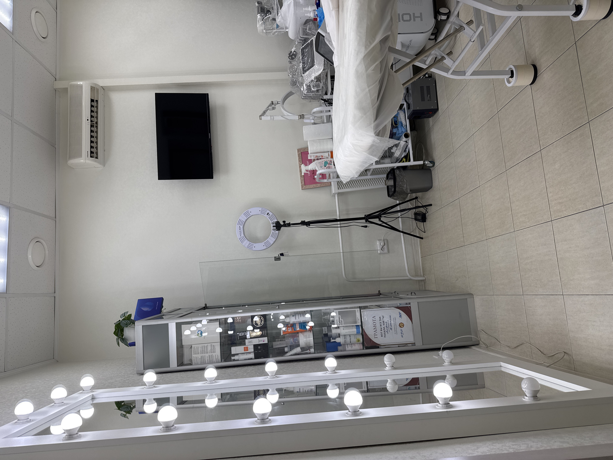 The image depicts an interior scene of what appears to be a beauty or cosmetic treatment room. There is a tiled floor and white walls. Key elements within the frame include a beauty bed covered with white fabric, various cosmetic devices and machinery, and an assortment of products displayed in a glass-fronted cabinet. A ring light on a tripod stand is positioned in the middle ground. In the foreground, there is a white vanity mirror with multiple light bulbs. A wall-mounted air conditioning unit and a television screen are visible on the left wall. A plant in a pot rests on top of the cabinet. A diploma or certificate is displayed within the glass cabinet. The precise location is unknown.