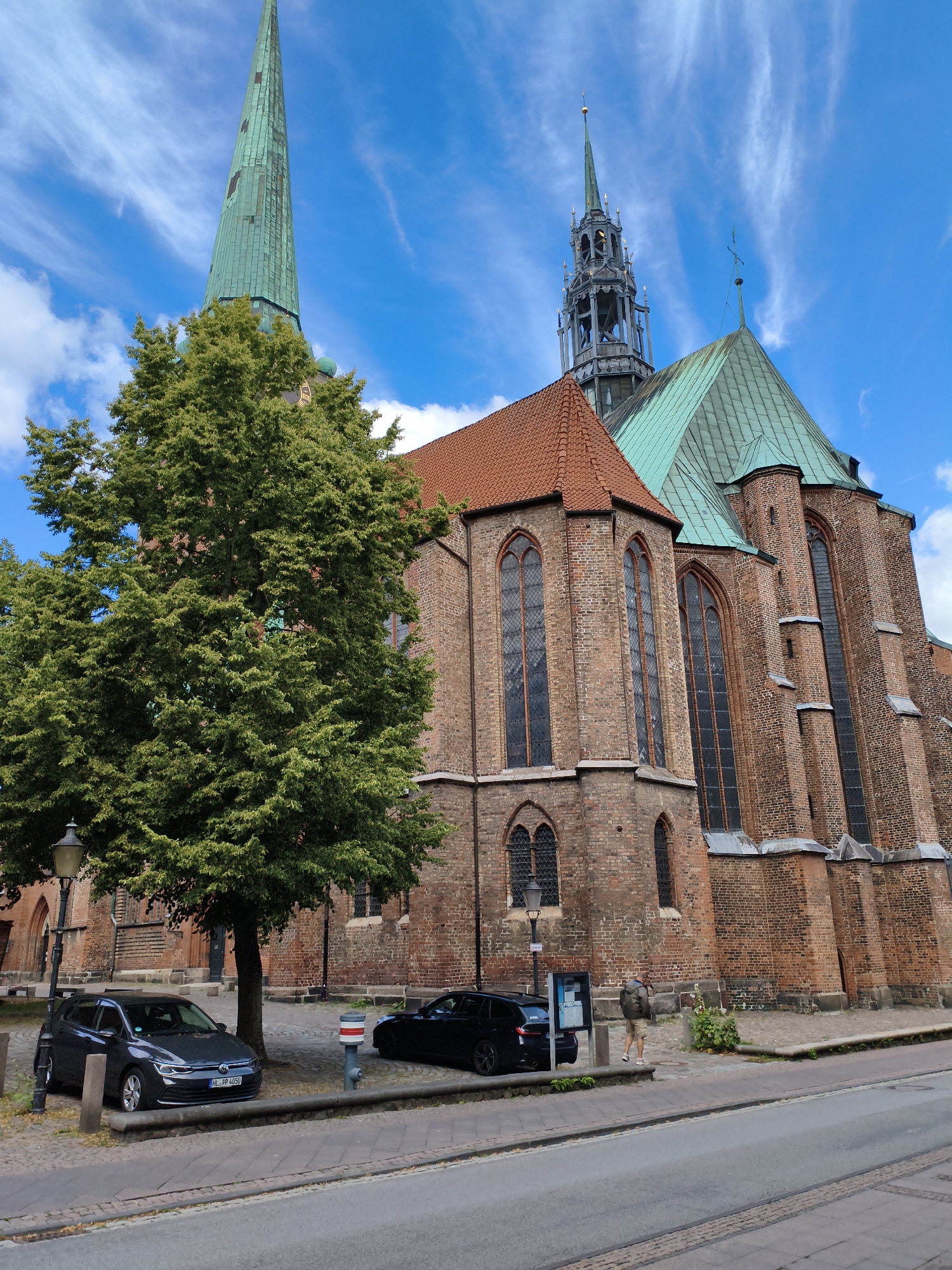 The image captures a street view of the Lübeck Cathedral in Lübeck, Germany. The cathedral, a prominent brick Gothic structure, features a tall green spire and a complex, darker spire structure to its right. Red-tiled roofs top sections of the cathedral, and large Gothic windows with dark panes are visible on the brick facade. A large, mature green tree with dense foliage stands in the foreground to the left of the cathedral, partially obscuring one of the spires. In front of the tree, a dark gray Volkswagen hatchback is parked on a cobblestone street. To the right of this car, another dark-colored sedan is parked. A man wearing a light brown jacket and shorts is walking away from the camera towards the right side of the frame, near the cathedral's entrance. The sky is blue with wispy white clouds. The street appears to be paved with asphalt, with a cobblestone sidewalk bordering it. A lamppost is visible next to the parked Volkswagen.