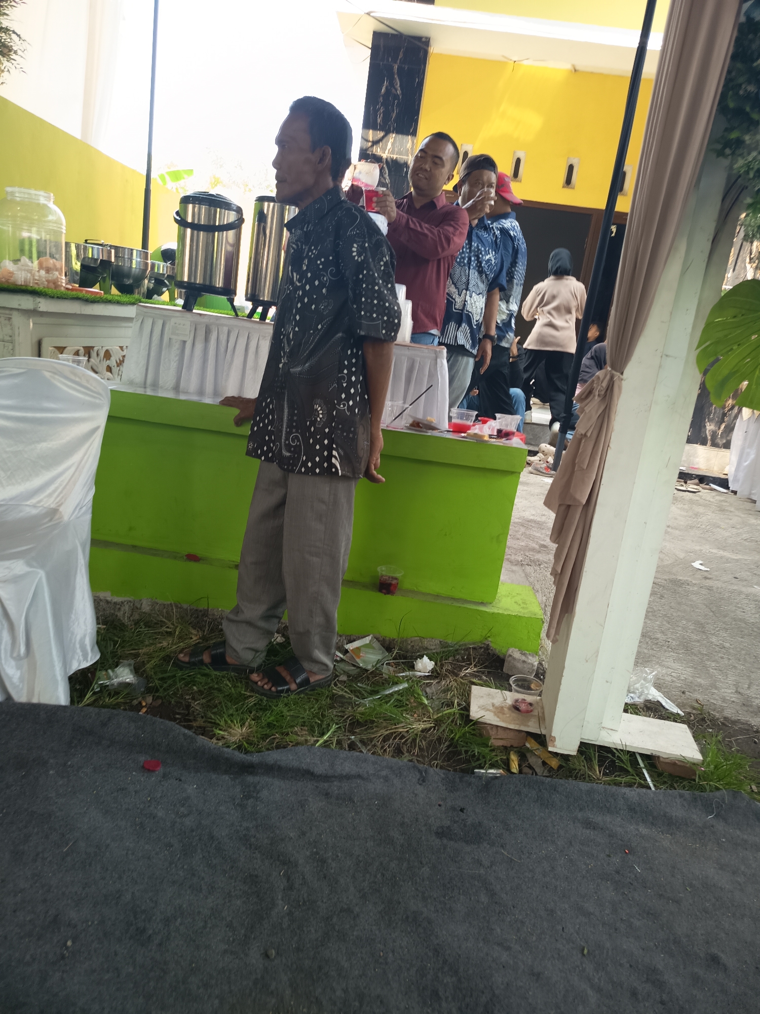 A medium shot captures an outdoor gathering with several people, a food or drink station, and draped fabric. In the foreground, a man with dark, short hair and a patterned black and white shirt stands facing left, his body angled towards the viewer. He wears grey pants and dark sandals. He appears to be looking towards the right, his mouth slightly open. Behind him, to his left, a table is set up with white draped cloth. On the table, there are large silver urns and a clear dispenser filled with ice and what appear to be pastries or bread rolls. To the right of the main subject, a few more people are visible. One man in a maroon shirt is holding a drink and looking towards the viewer. Another man in a blue and white patterned shirt is also holding a drink. Further back, a woman in a light brown top and dark pants is walking away from the camera. A large, tan-colored fabric is draped over a white pillar on the right side of the frame, partially obscuring the background. The ground is covered in grass and some debris. The scene suggests a casual event, possibly a catered gathering or celebration, set up outdoors.