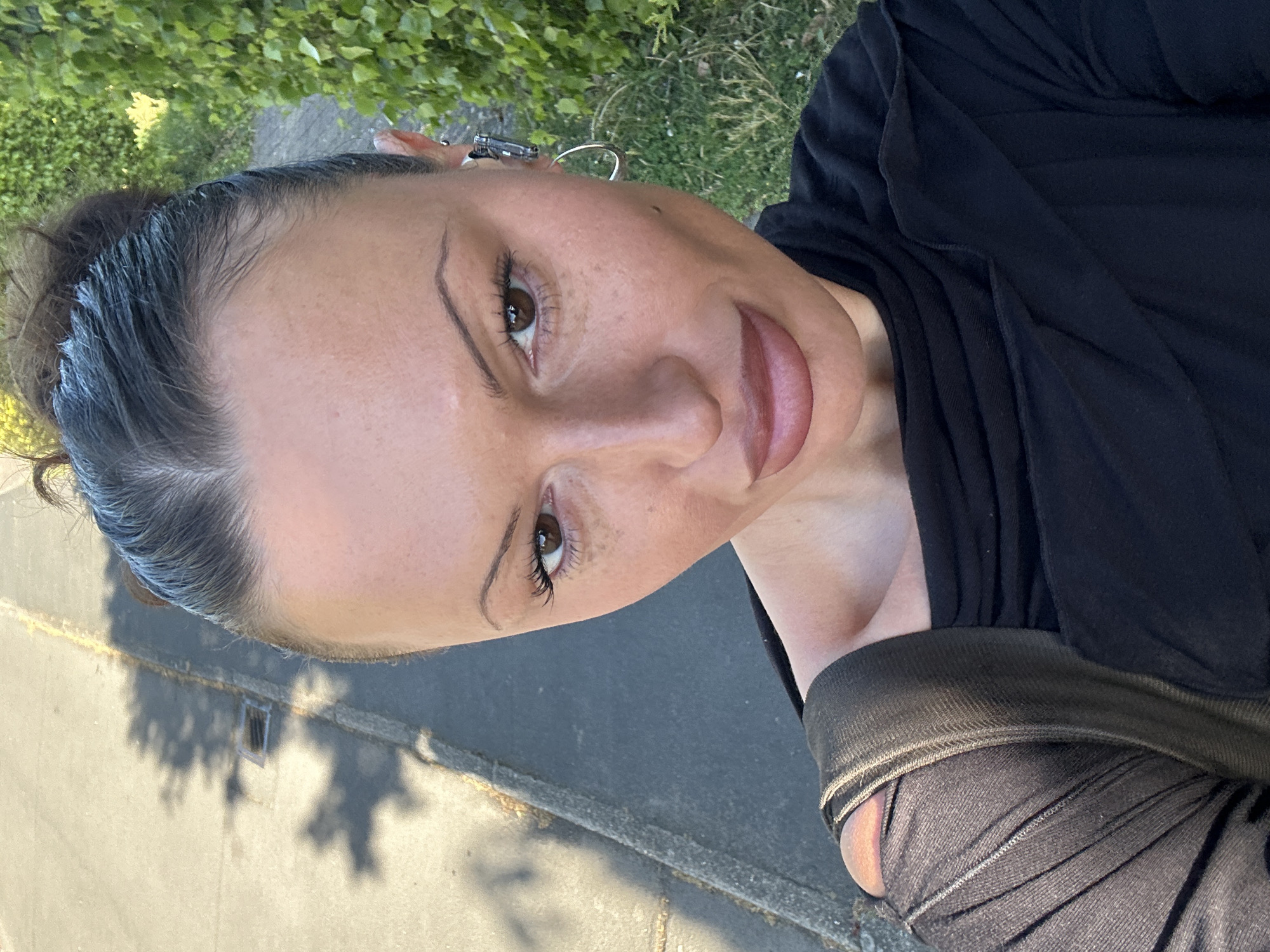 The image is a brightly lit, vertical outdoor selfie of a woman taken in Tunbridge Wells, United Kingdom, from an elevated, slightly angled perspective. The woman, with a medium skin tone and subtle freckles, looks up towards the camera with a soft, closed-lip smile, conveying a serene expression. Her dark brown hair is pulled back, with visible baby hairs around her forehead and some strands appearing grey or reflecting light. She has dark brown eyes, precisely defined eyebrows, and long eyelashes, enhanced with what appears to be eyeliner. Her full lips are colored with a natural pink/nude shade. She wears a black top or dress with visible pleats or gathers on the fabric. Distinctive earrings adorn her ears: one is a simple hoop, and the other is a more intricate, multi-component ear cuff.
The background features lush green foliage from trees or bushes. Below the foliage, a paved surface, likely a road or pathway, is visible, marked by long, distinct shadows from unseen trees, indicating either early morning or late afternoon sunlight on a clear day. A small, square grate or drain cover is faintly discernible on the paved ground. No text is visible in the image.