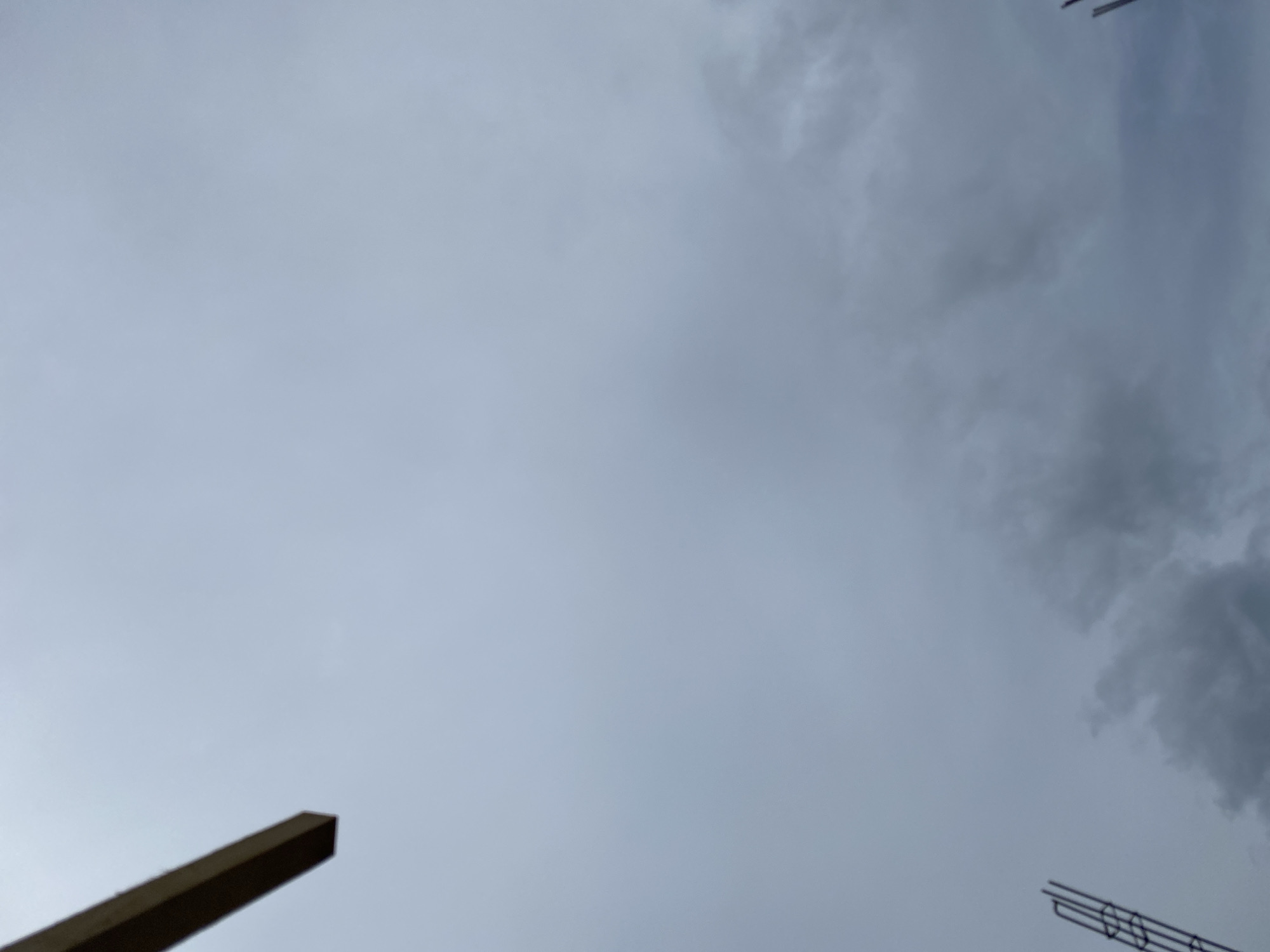 An overcast sky fills the frame of the image. In the upper left quadrant, a segment of a wooden beam extends diagonally from the top edge towards the center of the image. In the bottom left corner, a vertical construction element, possibly rebar or scaffolding, is visible. The sky itself is a uniform light gray, with subtle darker gray clouds appearing towards the bottom of the image. The scene suggests an outdoor environment, likely a construction site, under a cloudy sky. There are no people or discernible actions taking place in the image.
