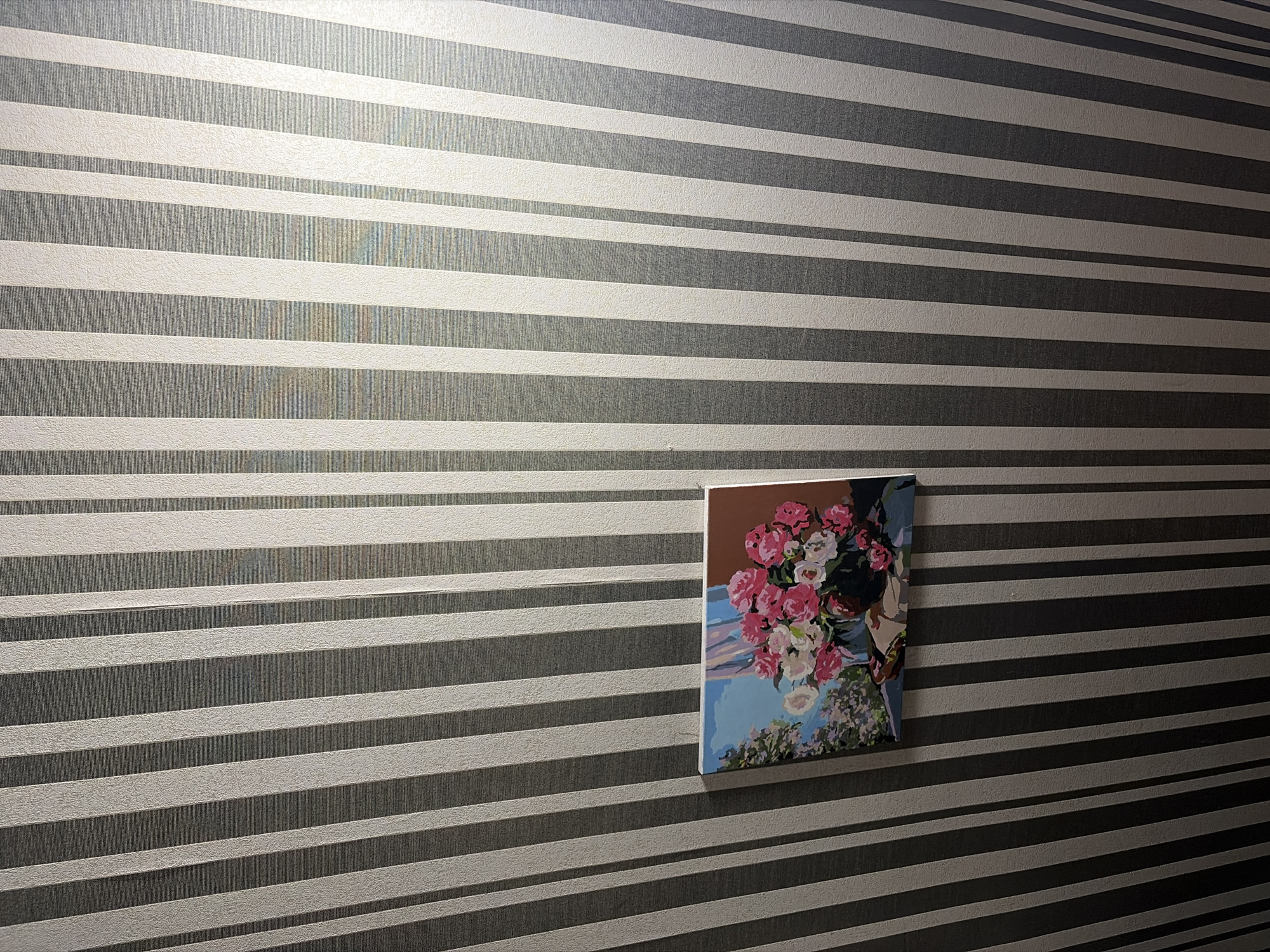 A framed painting of a bouquet of pink and white roses in a vase hangs on a striped wall. The wall has alternating vertical stripes of light gray and dark gray. The painting is positioned in the lower left quadrant of the image, leaning against the wall. The overall scene appears to be an indoor setting. There is no indication of the city of Sochi, Russia, or any specific location.
