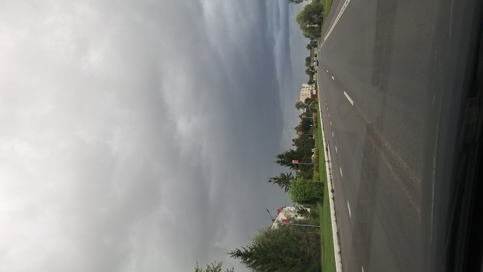 A view from a vehicle driving on a paved road towards buildings and trees under a cloudy sky. The road has white lane markings and a white curb. On the left side of the road, there are trees, a building, and several red flags on poles. On the right side of the road, there are trees and buildings visible in the distance. Two cars are on the road in the distance. The sky is overcast with dark clouds. This scene is in the city of Myadzyel, Belarus.