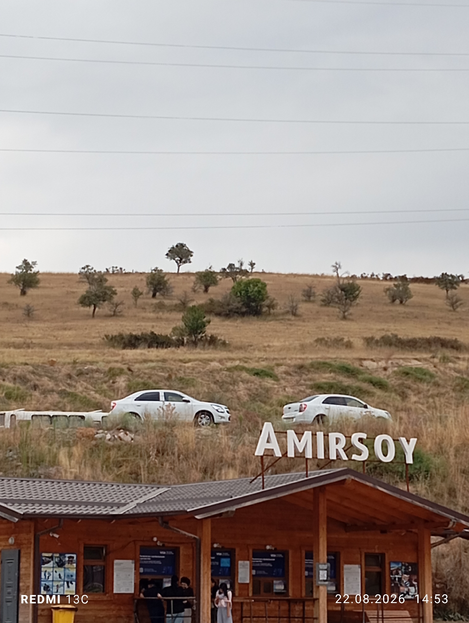 The image depicts a landscape scene in Amirsay, Uzbekistan, featuring a prominent wooden structure at the forefront, likely a building or kiosk. The structure has a sloped, dark-tiled roof supported by wooden beams. Several windows are visible on the front facade of the building, some displaying posters or information. A few individuals are gathered near the windows. To the left of the building, the text "REDMI 13C" is visible, indicating the phone used to capture the image, and the date "22.08.2026 14:53" is displayed in the bottom right corner.
Above the wooden structure, a large white sign spells out "AMIRSOY". In the middle ground, a dry, grassy hillside is visible, dotted with scattered trees and bushes. Two white cars are parked on the slope. The car on the left is partially obscured by a concrete structure. The car on the right is clearly visible. The background consists of a wide, undulating grassy landscape under an overcast sky. Power lines are stretched across the upper portion of the image, running horizontally.
