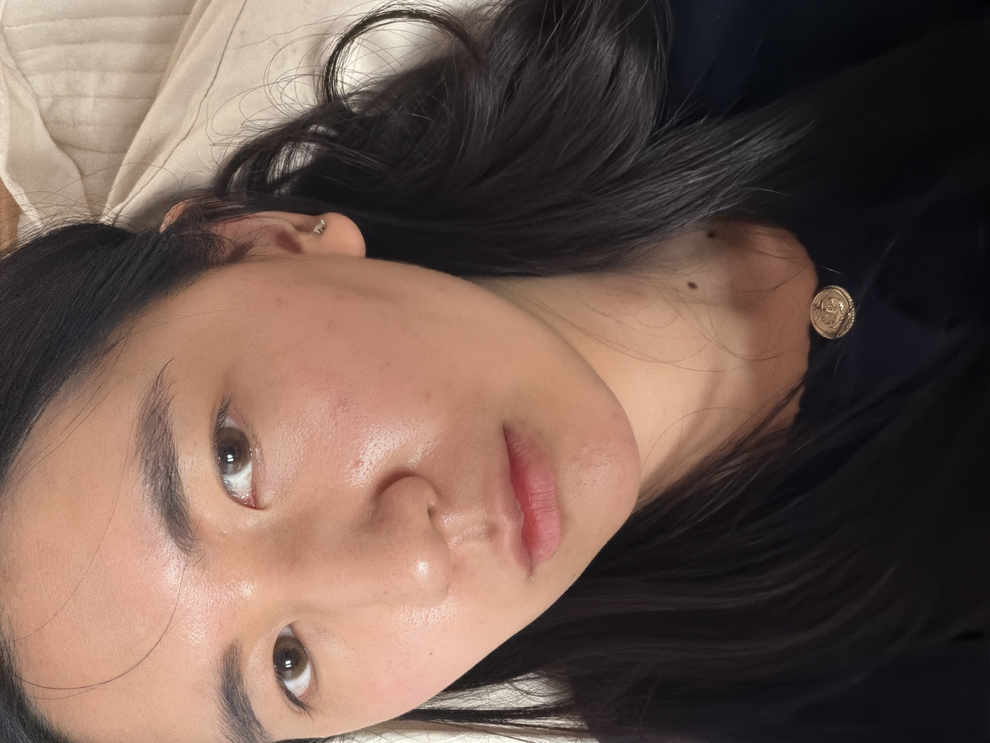 The image shows a young woman lying down, possibly in bed. She has dark hair and is wearing a black top. She is fair-skinned with brown eyes and is wearing an earring in her left ear and a round, possibly gold, earring on the opposite side. The background consists of a cream-colored surface, possibly a pillow or bedding. The location is not identifiable as Paris, France.
