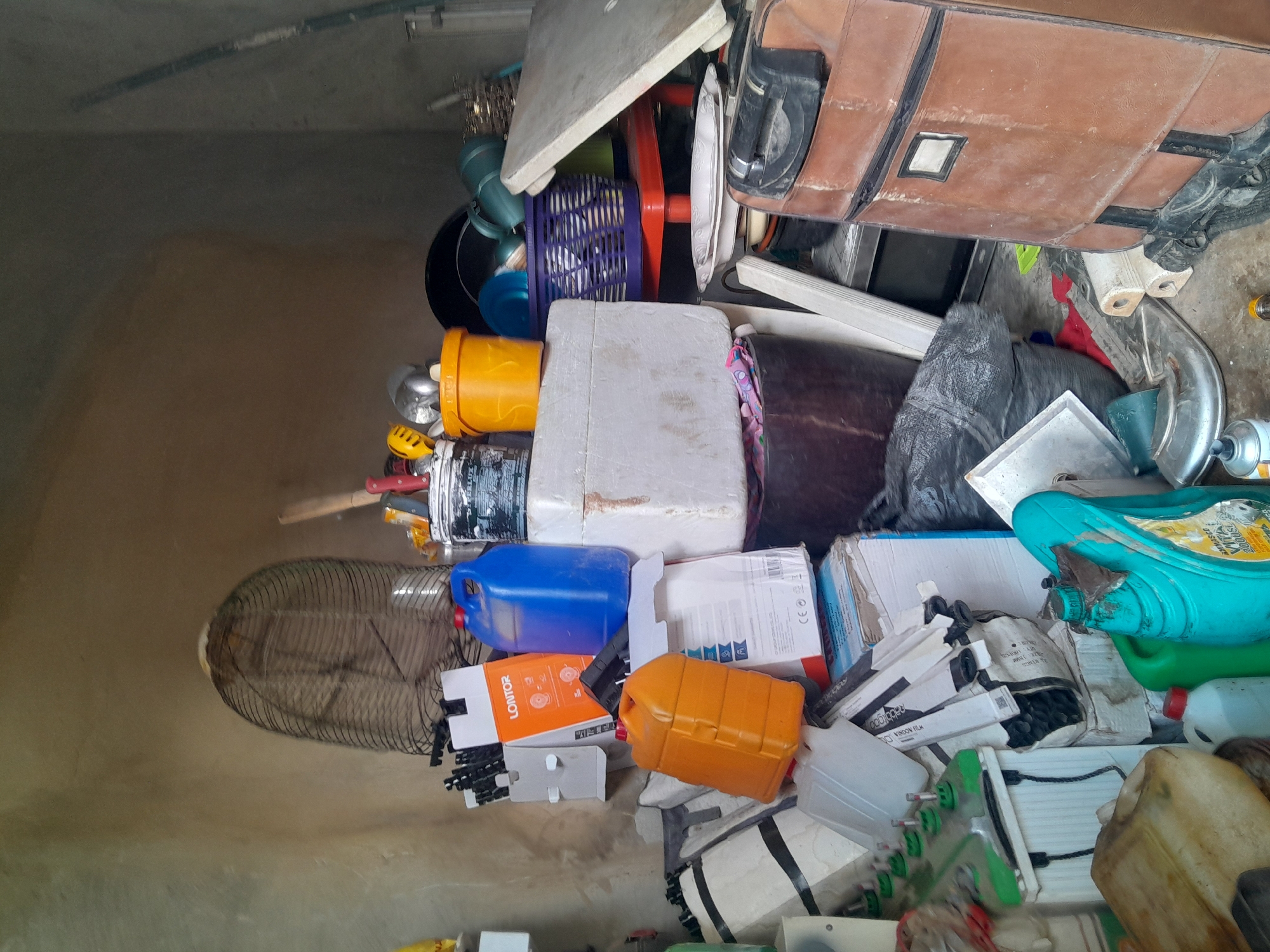 In Gauraka, Nigeria, a cluttered room contains various items piled high. A large, dark brown suitcase sits on the right side of the frame. To its left, a dark grey trash bag is partially visible. Several white and light grey boxes are stacked together, with some appearing to be storage containers.
On a lower level, a light brown plastic jerrycan is present, as is a smaller white plastic bottle with a red cap. A bright blue plastic container with a black handle is positioned on top of other items. An orange plastic jerrycan is also stacked amongst the boxes.
In the background, a large, dark wire cage or fan guard is positioned against a textured, beige wall. Various tools, including what appear to be knives and a hammer, are visible near the top of the frame. Other assorted objects, including buckets, baskets, and what looks like a white board or panel, are scattered throughout the scene, suggesting a storage or workshop area. The lighting is somewhat dim, and the overall impression is one of disarray and accumulation of goods.