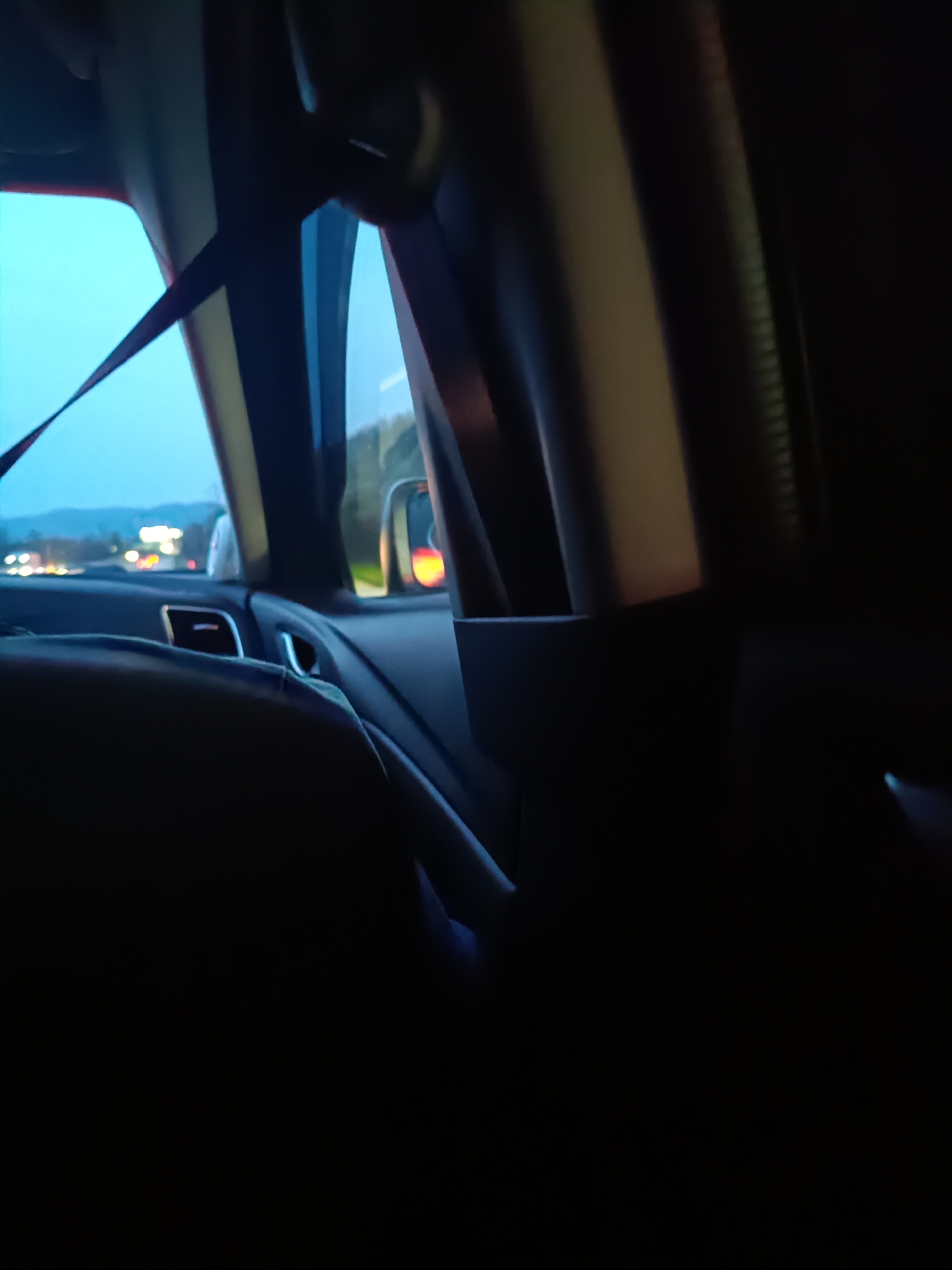 The image depicts the interior of a vehicle, viewed from the back seat. The forward view through the side window shows a road with oncoming traffic, indicating a nighttime or dusk setting. A seatbelt is visible across the window. The side mirror reflects a red light, likely from another vehicle. The general environment outside appears to be a suburban or semi-rural area with trees and hills in the distance.