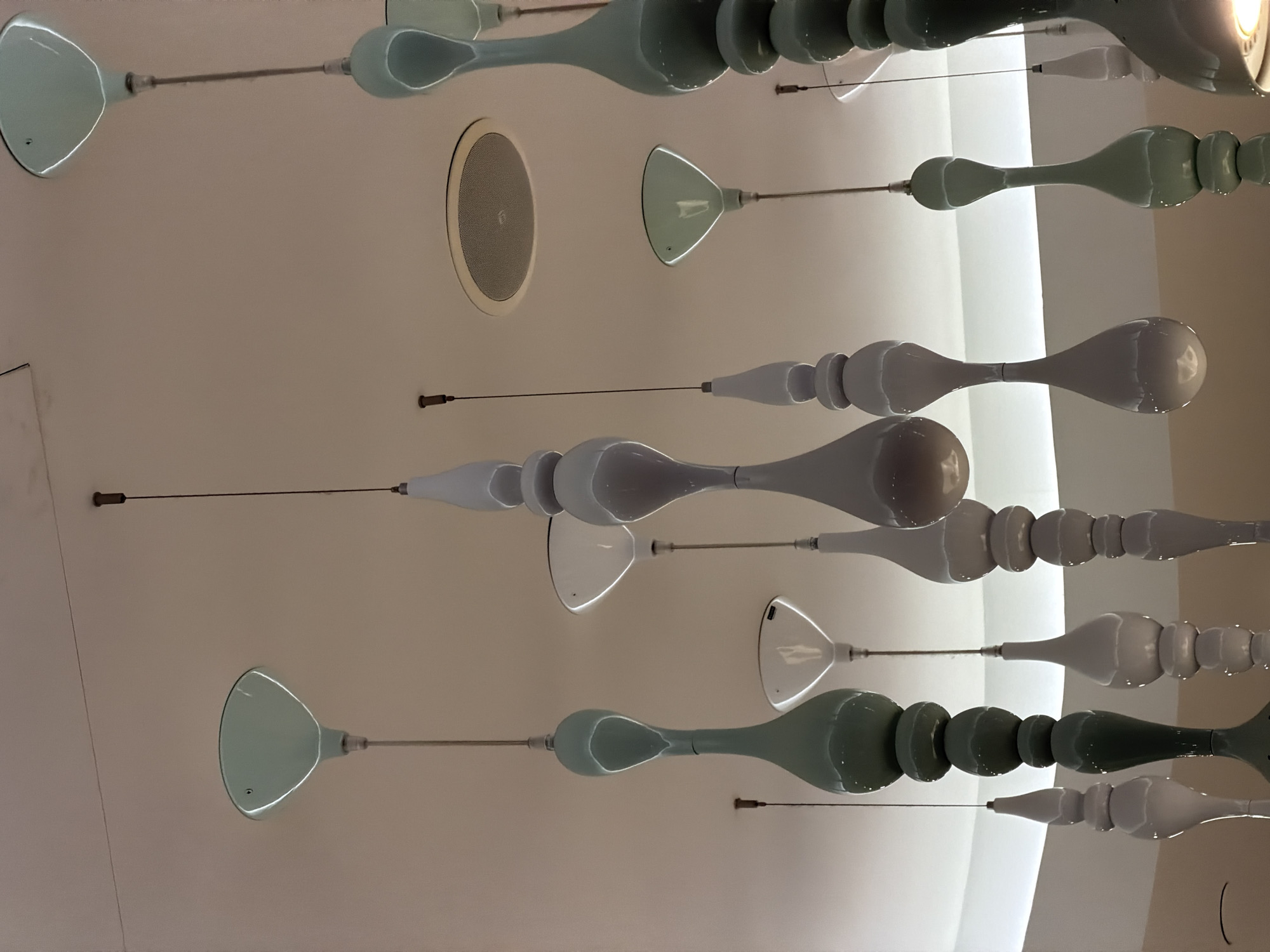 The image shows a ceiling with an array of decorative, elongated fixtures suspended from it. These fixtures consist of a rounded, bowl-shaped component attached to a thin rod that connects to a series of stacked, bulbous elements. The color scheme is primarily muted greens and whites, with a glossy finish suggesting that the fixtures are made of glass or a similar reflective material. The arrangement is seemingly random and covers a significant portion of the ceiling. In addition to the fixtures, there is a circular speaker embedded in the ceiling. The background is a pale, off-white surface with a subtle gradient, indicating light emanating from a nearby source. Based on the style of the fixtures and architecture, it's possible this is an artistic installation located in Moscow, Russia, but further contextual information is needed for confirmation.
