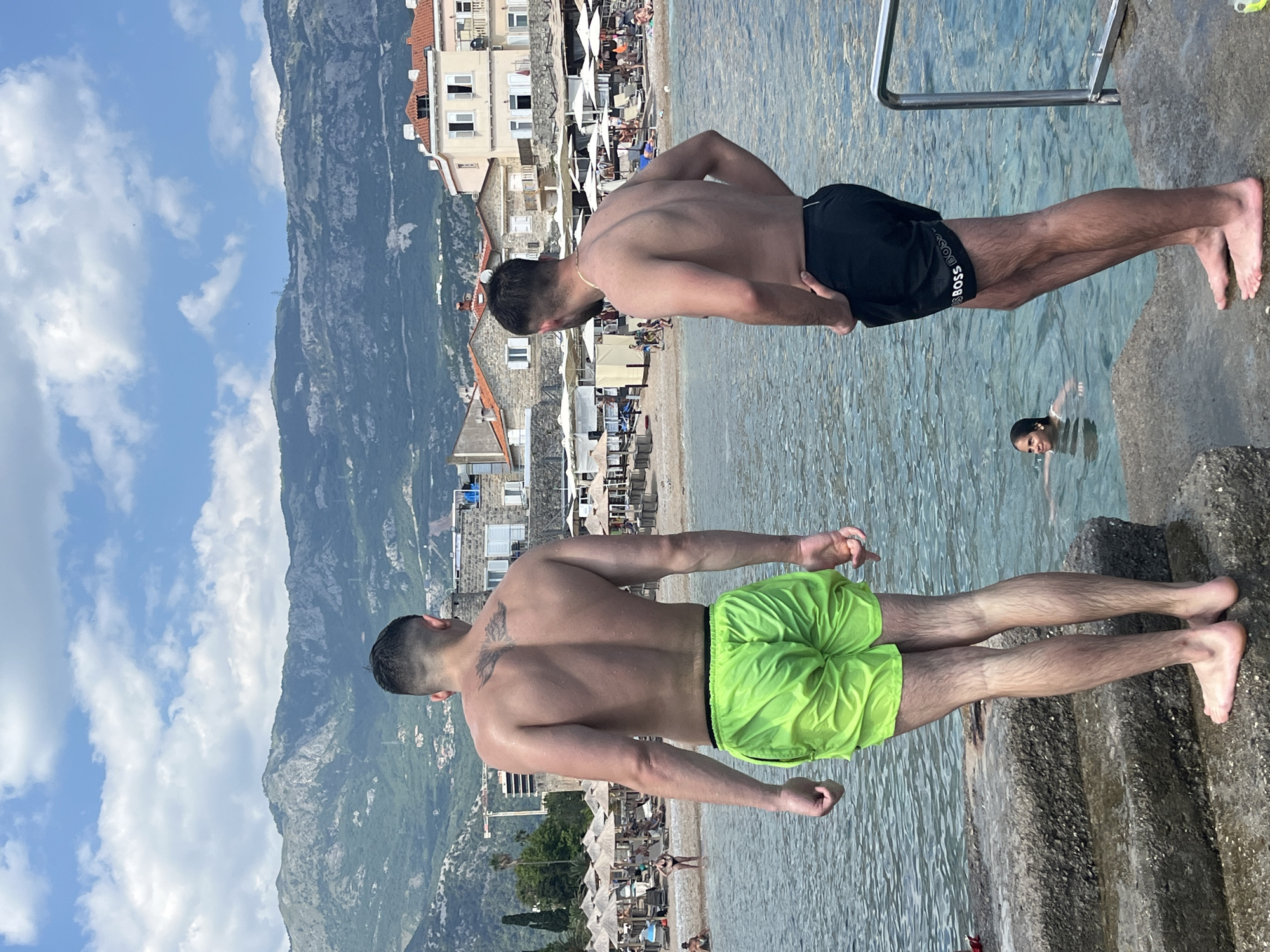 Two men stand on a concrete embankment at the edge of the sea. The man on the left is facing away from the camera, wearing bright green swim shorts. The man on the right is also facing away, wearing black swim shorts with "BOSS" written on the side, and has his left hand on his hip. In the background, a beach is visible with numerous umbrellas and lounge chairs. Beyond the beach, buildings with red-tiled roofs are clustered together. In the far background, a large, green, mountainous landscape rises under a blue sky with scattered white clouds. A woman is in the water to the left of the man on the right, swimming. The scene is located in the city of Budva, Montenegro.
