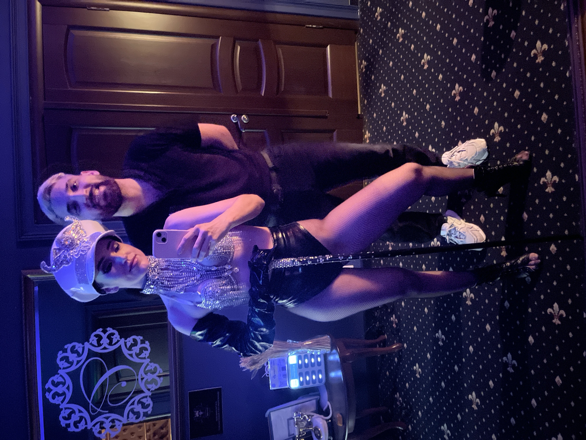 The image depicts two individuals indoors, illuminated by deep blue and purple ambient lighting. On the left, a woman stands in a performance-style costume, holding a light-colored smartphone (likely an iPhone) to take a selfie. She wears a white military-style cap adorned with ornate silver embellishments, a silver beaded halter top exposing her midriff, shiny black long gloves extending above the elbow, black patent leather high-waisted shorts, and fishnet stockings leading to dark open-toed ankle boots. She holds a black, possibly rhinestone-accented, cane or pole in her left hand.
Standing behind and slightly to her right is a man with light-colored hair and a beard, smiling towards the camera. He is dressed in a black short-sleeved polo shirt, dark pants, and white athletic shoes with light soles.
The background features a dark wall. On the left, a large, ornate framed mirror reflects a stylized white "P" monogram within a decorative scrollwork border. To the left of the woman, a small dark wooden side table holds a vintage-style white rotary telephone, a modern white charging station with multiple glowing blue ports, and a vase containing dried reeds or grasses. On the right, two dark wooden panel doors with a silver handle are visible. The floor is covered with a dark blue or black carpet patterned with repeating light-colored fleur-de-lis motifs and small diamonds.
Based on the distinctive "P" monogram and carpet pattern, the location is identified as the P.S. Hotel in Kazan, Russia.