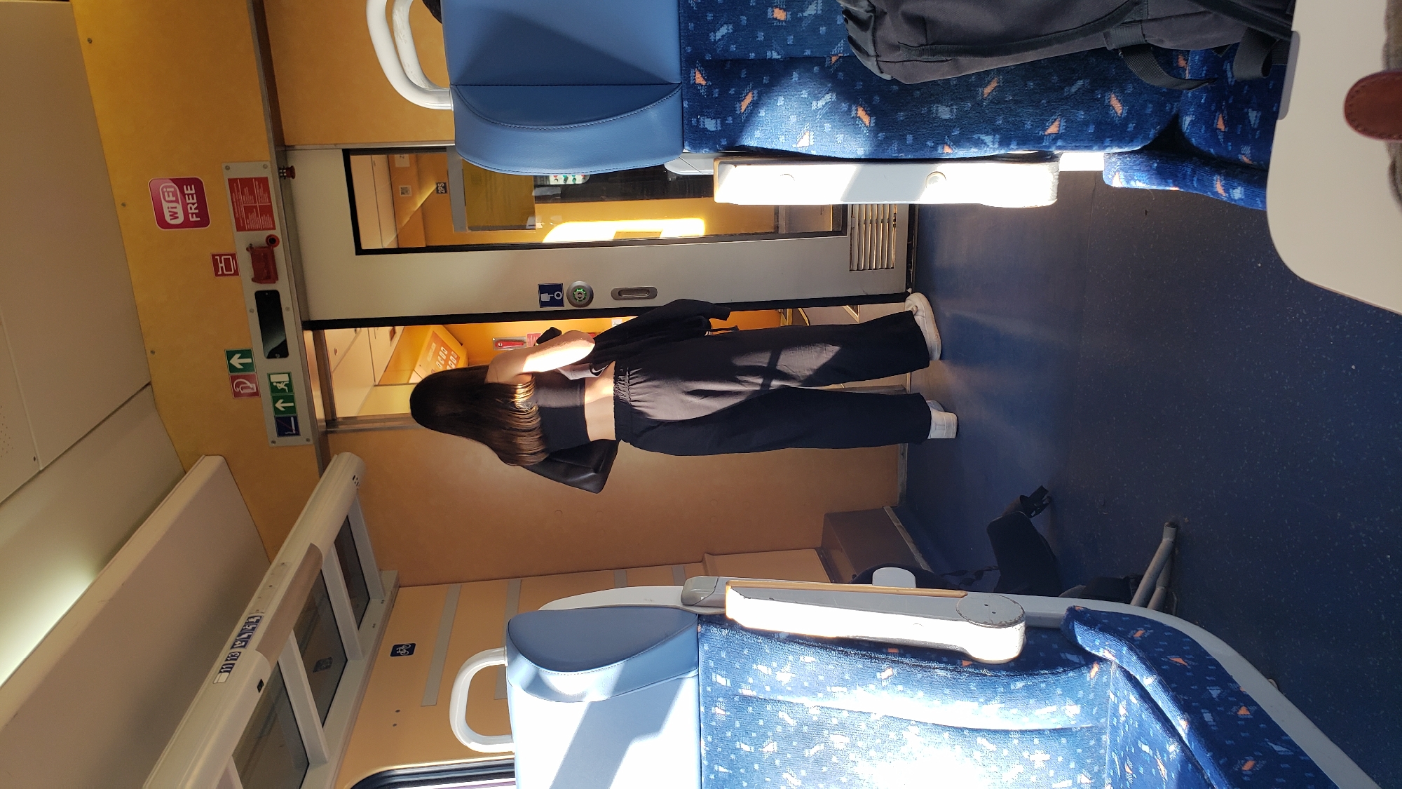 The image depicts the interior of a train car. In the foreground, blue seats with a patterned design are visible. A young woman with long dark hair is standing in the aisle, facing away from the camera and towards a doorway. She is wearing a black crop top and black wide-leg pants, with white sneakers. She is holding a black jacket over her left shoulder. The train car has yellow walls and overhead lighting. Signs are visible on the wall, including a "WIFI FREE" sign and directional arrows. Sunlight is streaming in through the windows, casting long shadows. The floor of the train car is dark blue.