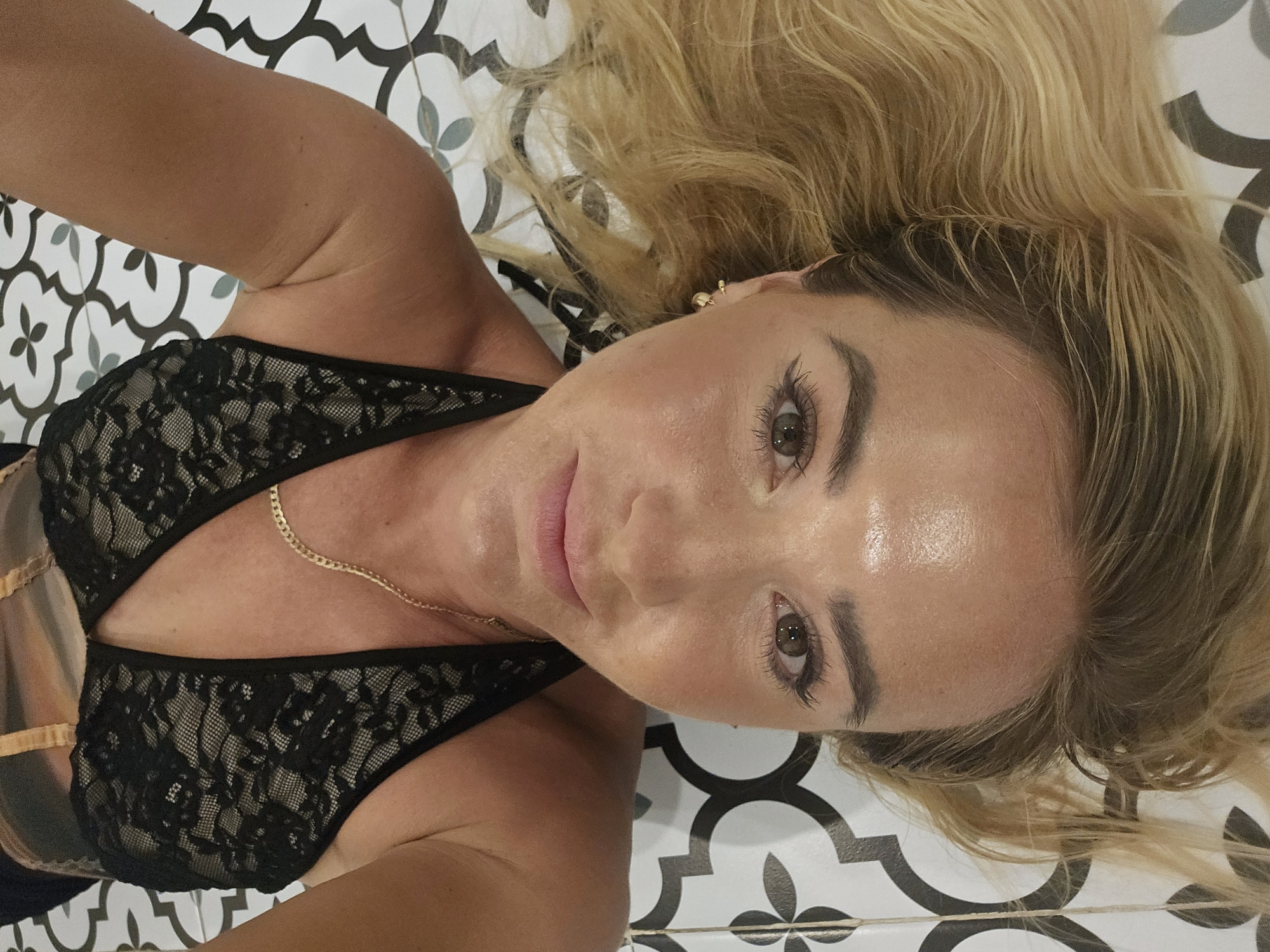 An upside-down, close-up, waist-up, selfie photograph shows a woman with blonde hair, lying on her back on a patterned tiled floor. The woman is wearing a black lace bra, a gold chain necklace, and a gold earring. Her face is the most prominent subject in the image, with her eyes open and looking towards the camera. Her makeup is visible, including eyeliner and mascara. The background consists of black and white patterned tiles, possibly in an indoor setting. There is no indication of Phu Thuy Ward, Vietnam.